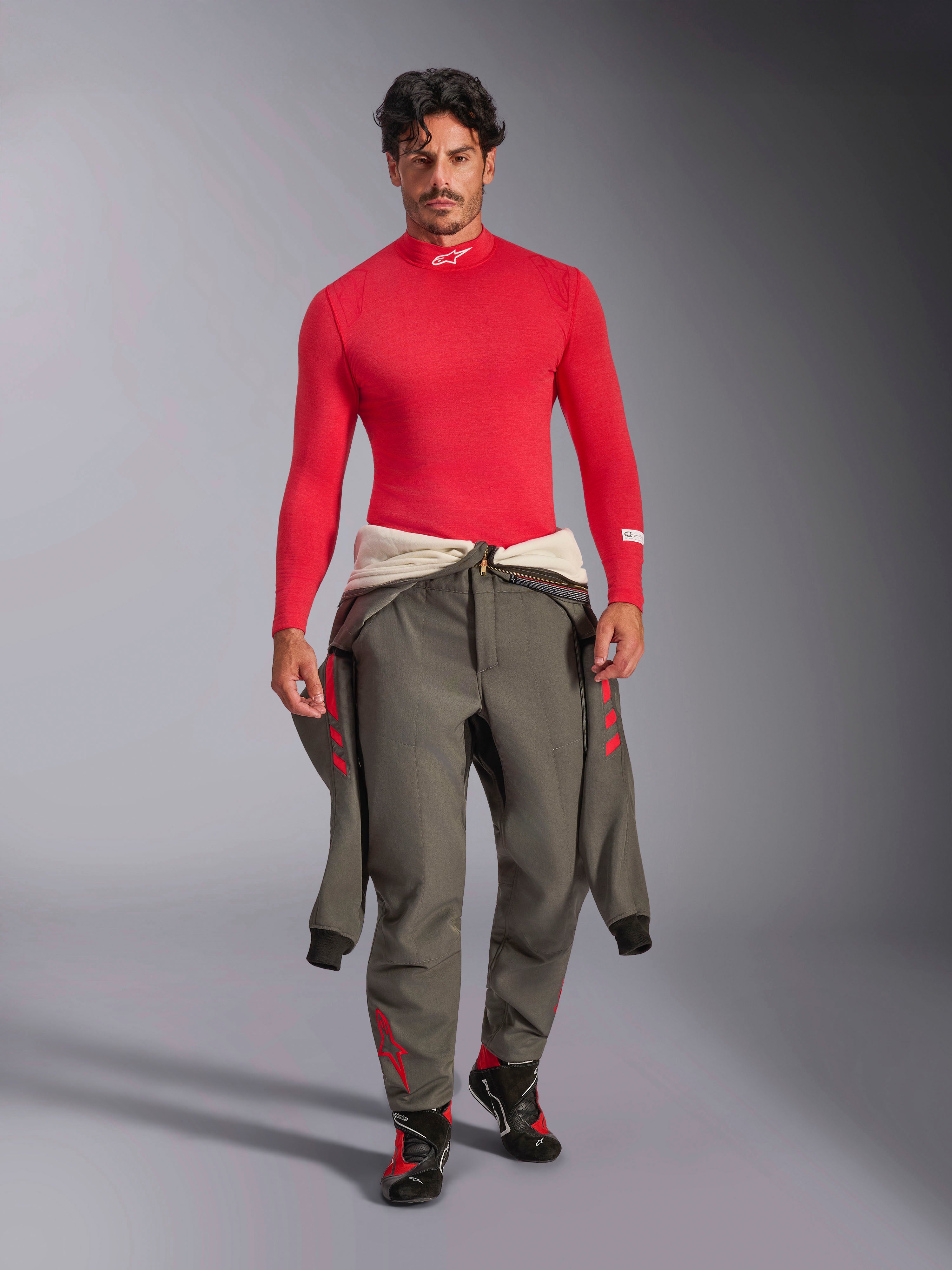 Maglia a maniche lunghe Alpinestars ZX Evo V3, sottotuta auto rosso, indossata da un modello con tuta da corsa grigia legata in vita e scarpe da pilota, collo a lupetto con logo Alpinestars bianco, abbigliamento professionale per motorsport leggero e resistente alle fiamme.