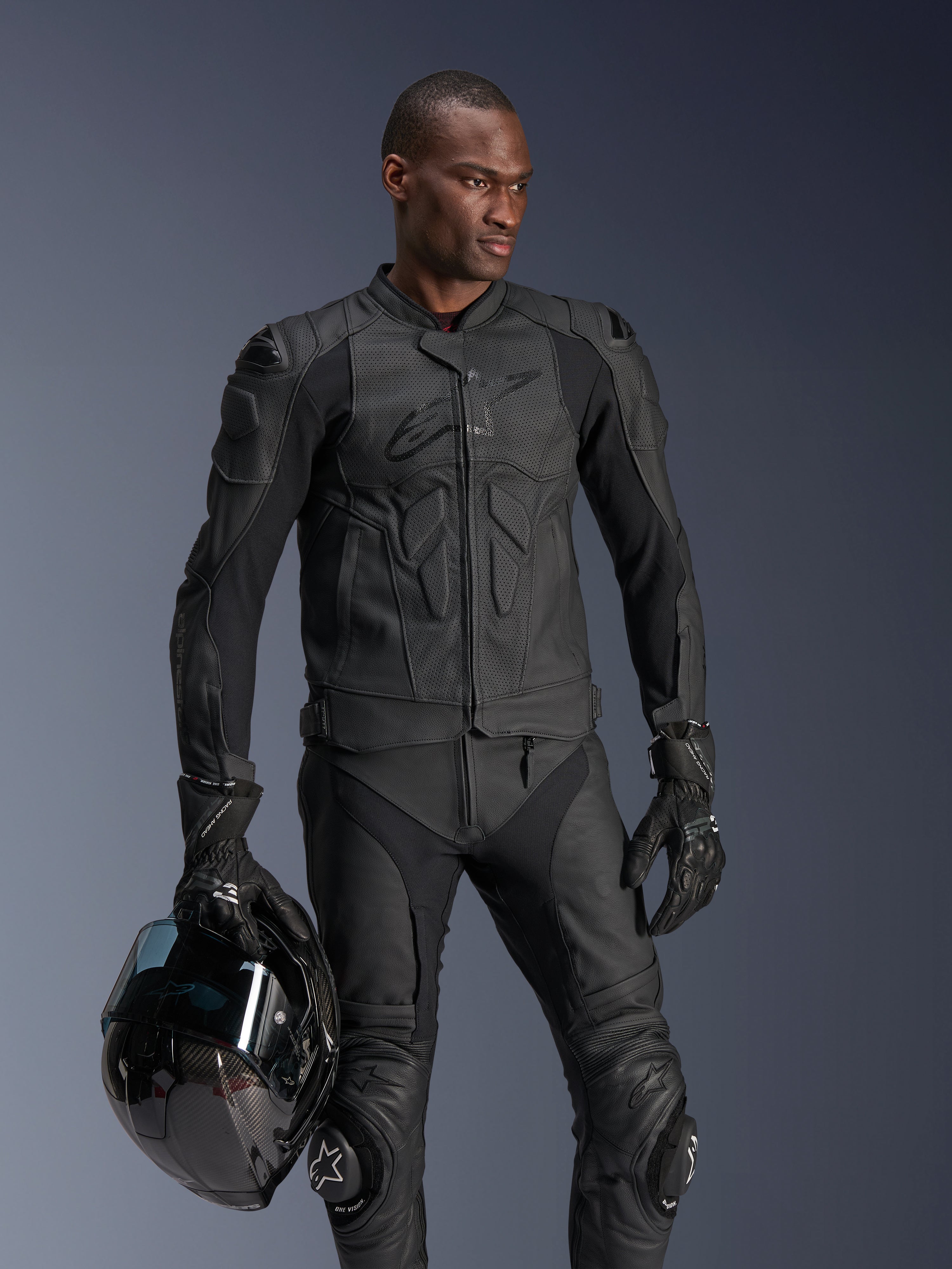 Giacca in pelle Alpinestars Dusk Airflow, nera, indossata da un pilota uomo con pantaloni in pelle e guanti coordinati, con in mano un casco nero, dotata di pannelli in pelle traforata e protezioni integrate per la guida sportiva in moto.