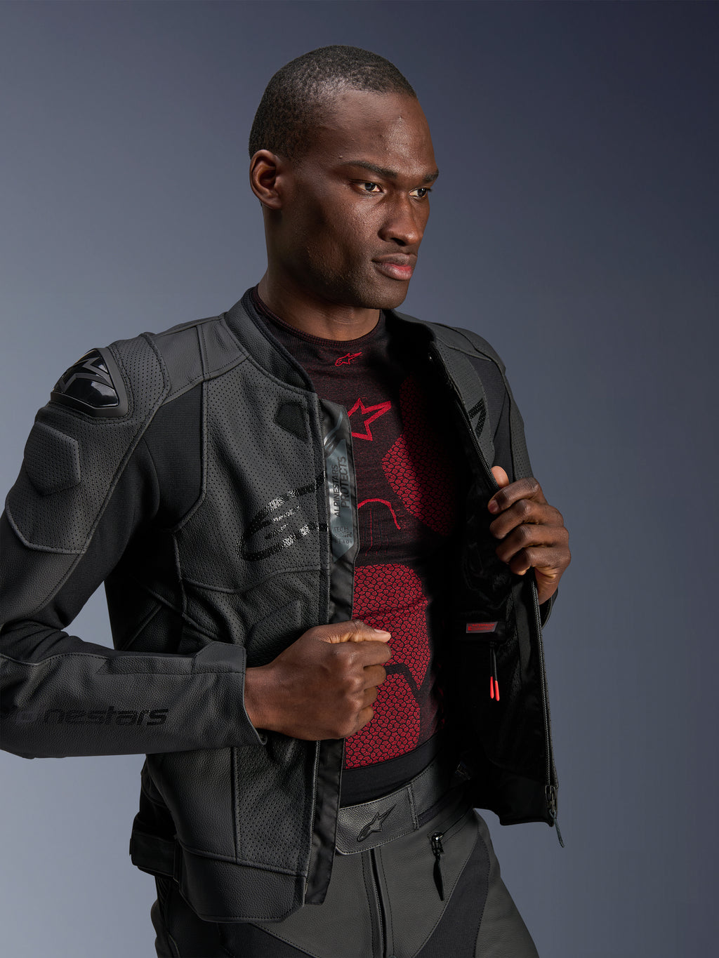 Giacca in pelle Alpinestars Dusk Airflow, colore Black Black, indossata da un modello sopra un base layer rosso e nero, con pannelli in pelle traforata, slider esterni sulle spalle DFS e vestibilità sportiva.