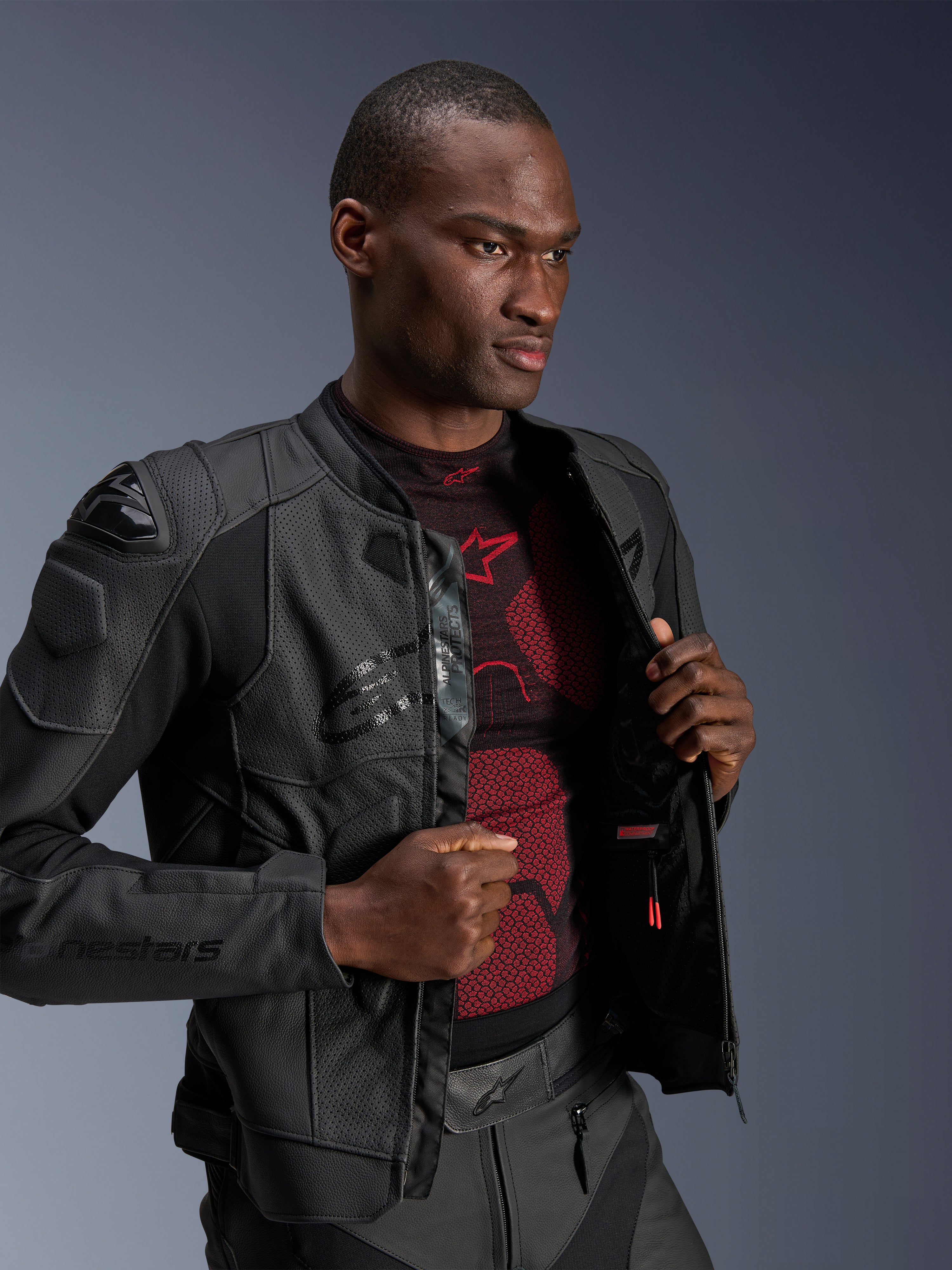 Giacca in pelle Alpinestars Dusk Airflow, colore Black Black, indossata da un modello sopra un base layer rosso e nero, con pannelli in pelle traforata, slider esterni sulle spalle DFS e vestibilità sportiva.