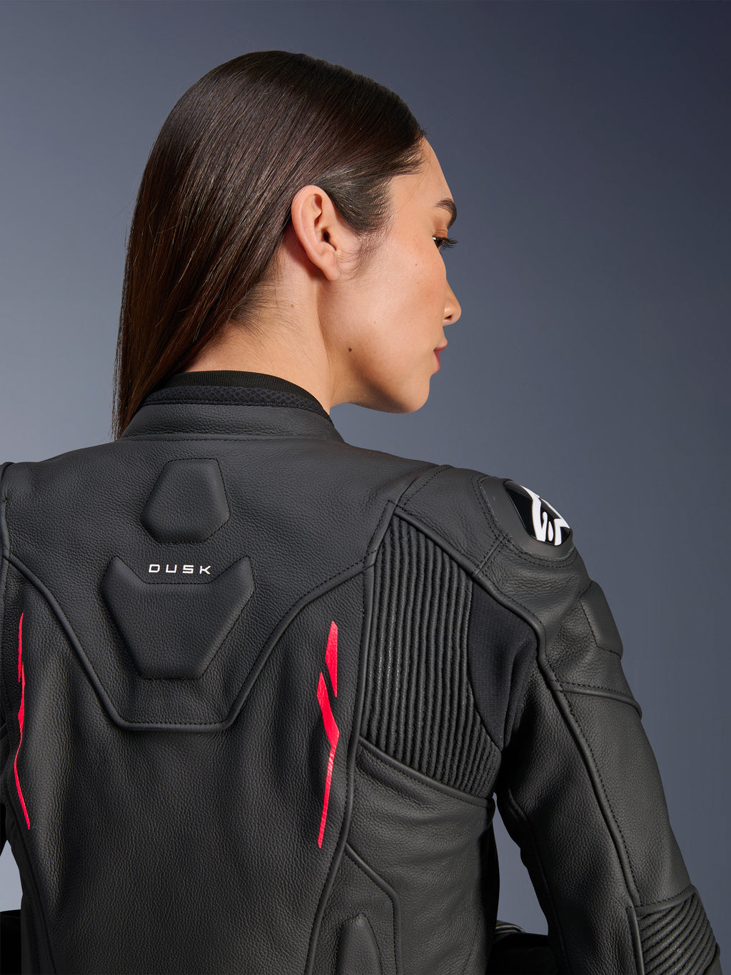 Donna che indossa la giacca in pelle Alpinestars Stella Dusk, pelle nera con accenti Diva Pink e logo bianco sulla spalla, vista posteriore con marchio DUSK e pannelli elasticizzati a fisarmonica, design ergonomico sportivo