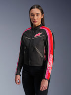 Donna che indossa la giacca Alpinestars Stella T-Dyno WR in Black Bright Red Fuchsia, giacca da moto in tessuto con una vestibilità da biker d'ispirazione vintage, caratterizzata da vivaci strisce fucsia e rosse sulle maniche e un logo sul petto, abbinata a pantaloni neri.