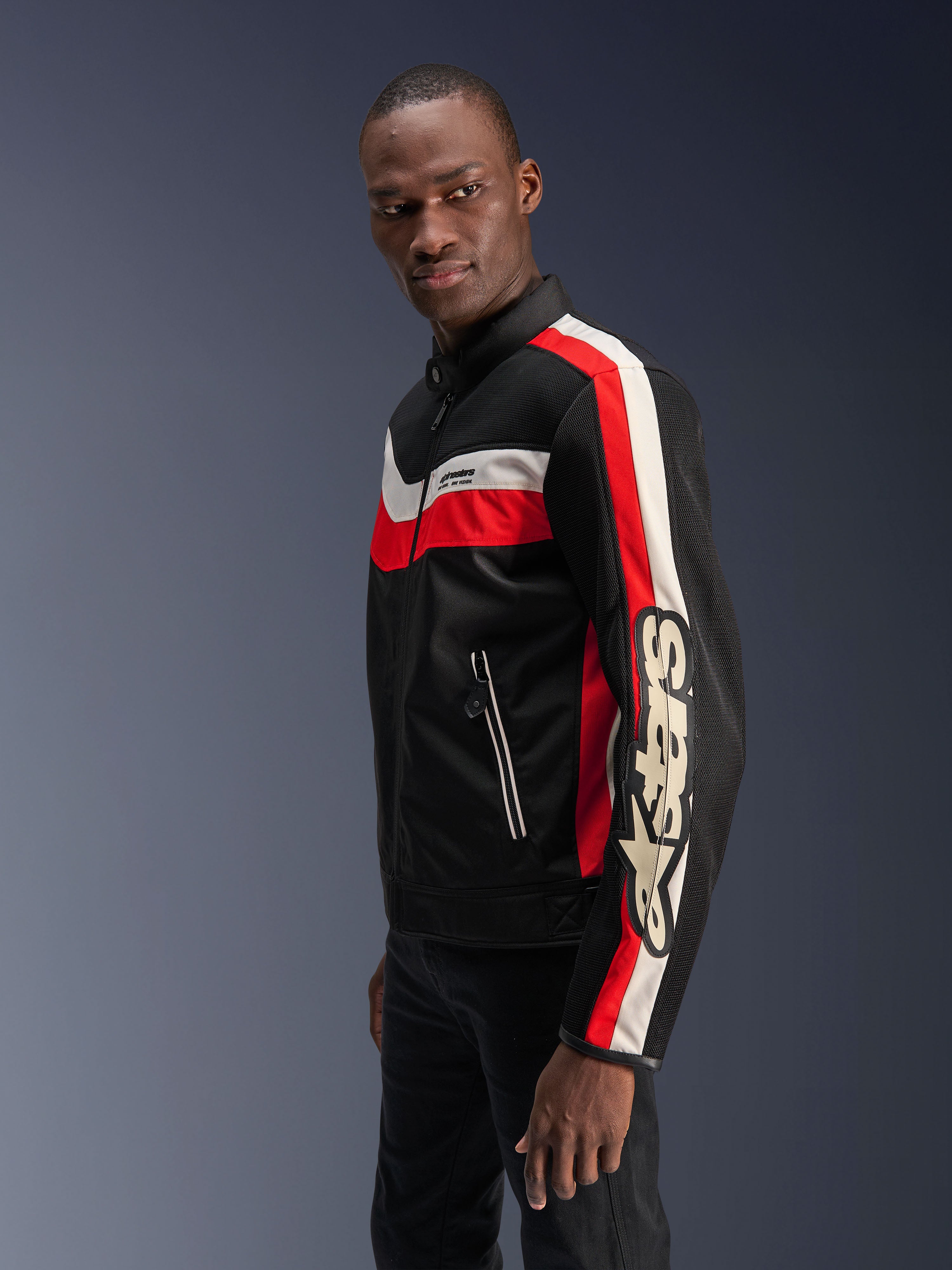 Modello uomo che indossa la giacca Alpinestars T-Dyno Air in nero, rosso acceso ed ecru, giacca da moto in tessuto d'ispirazione vintage con pannelli in mesh 3D, audaci strisce rosse e bianco sporco e marchio Alpinestars sulla manica.