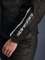 Giacca Alpinestars Stella T-SPS Air V2, giacca da moto da donna, nero e bianco, primo piano della manica con logo Alpinestars bianco e scritta T-SPS AIR, costruzione in mesh e tessuto per il flusso d'aria, indossata con pantaloni da moto neri