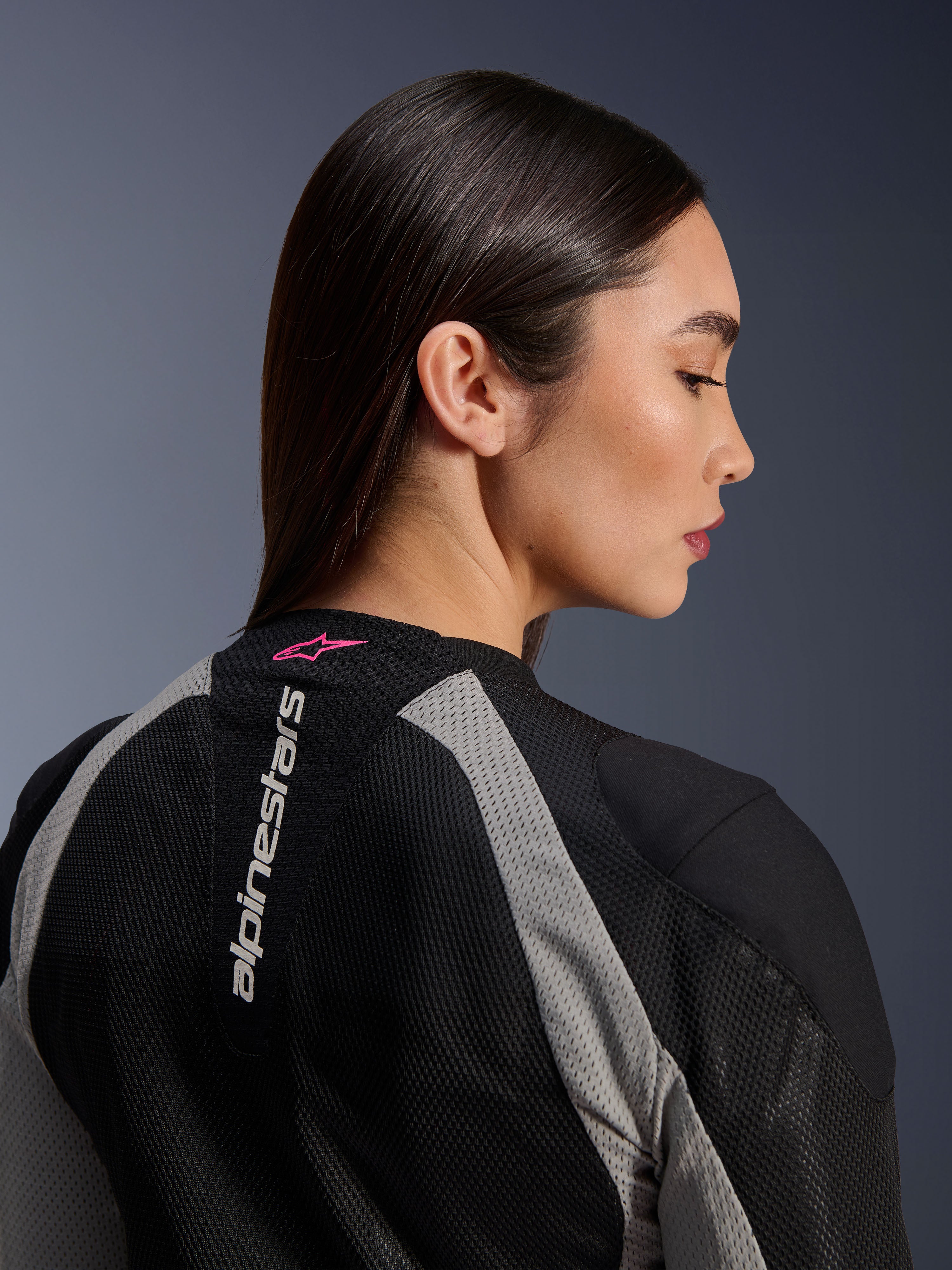 Profilo in primo piano di una donna che indossa la giacca Alpinestars Stella Troop-Air, costruzione in mesh nero e grigio Ash Gray, logo Alpinestars bianco verticale sulla parte superiore della schiena con logo a stella Pink Fluo, abbigliamento tecnico da moto leggero per donna