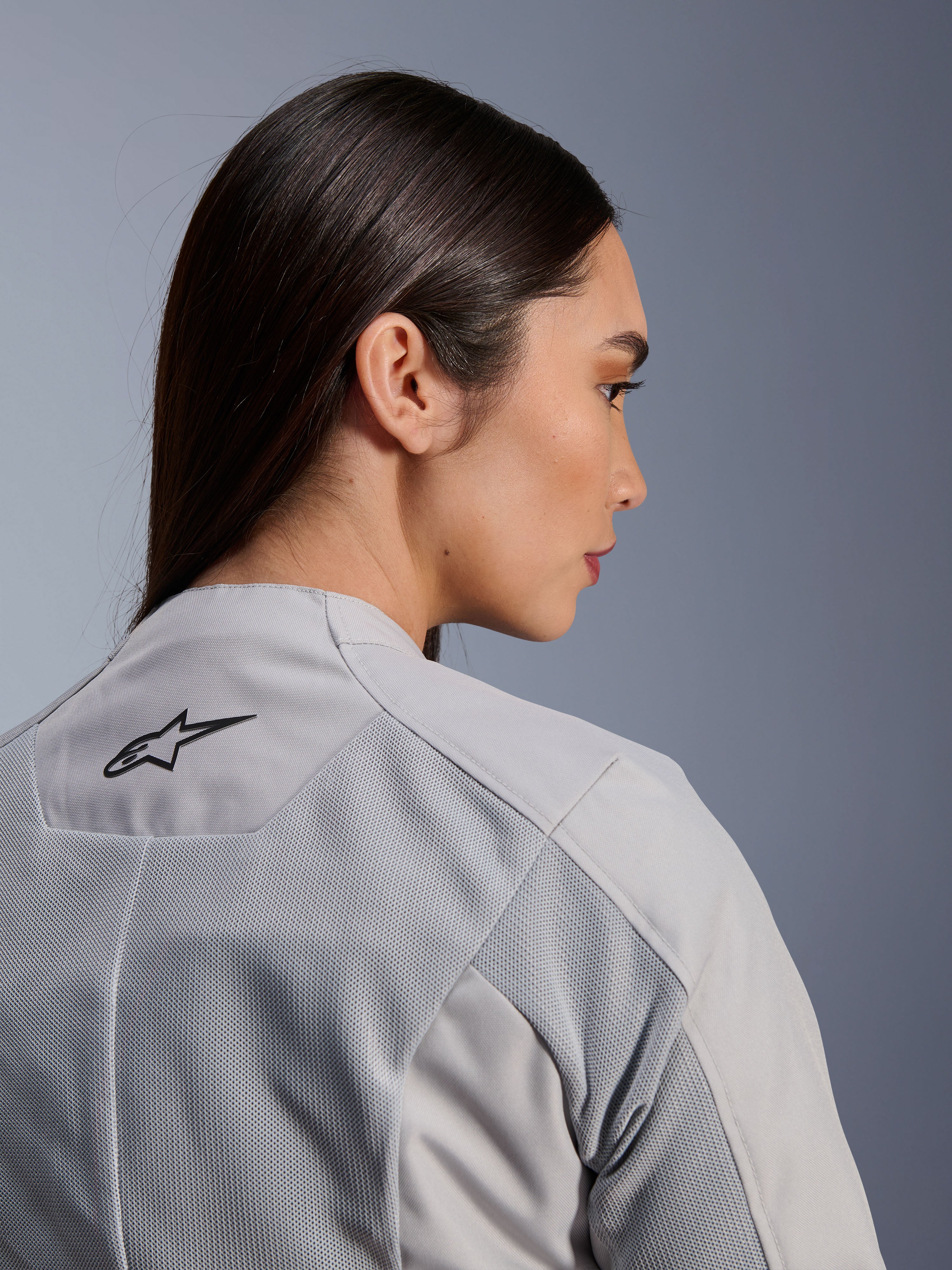 Donna che indossa la giacca Alpinestars Stella C-1 Air in argento, vista ravvicinata laterale-posteriore che mostra il logo Alpinestars nero, pannelli di ventilazione in rete tecnica e un design motociclistico urbano pulito e contemporaneo.