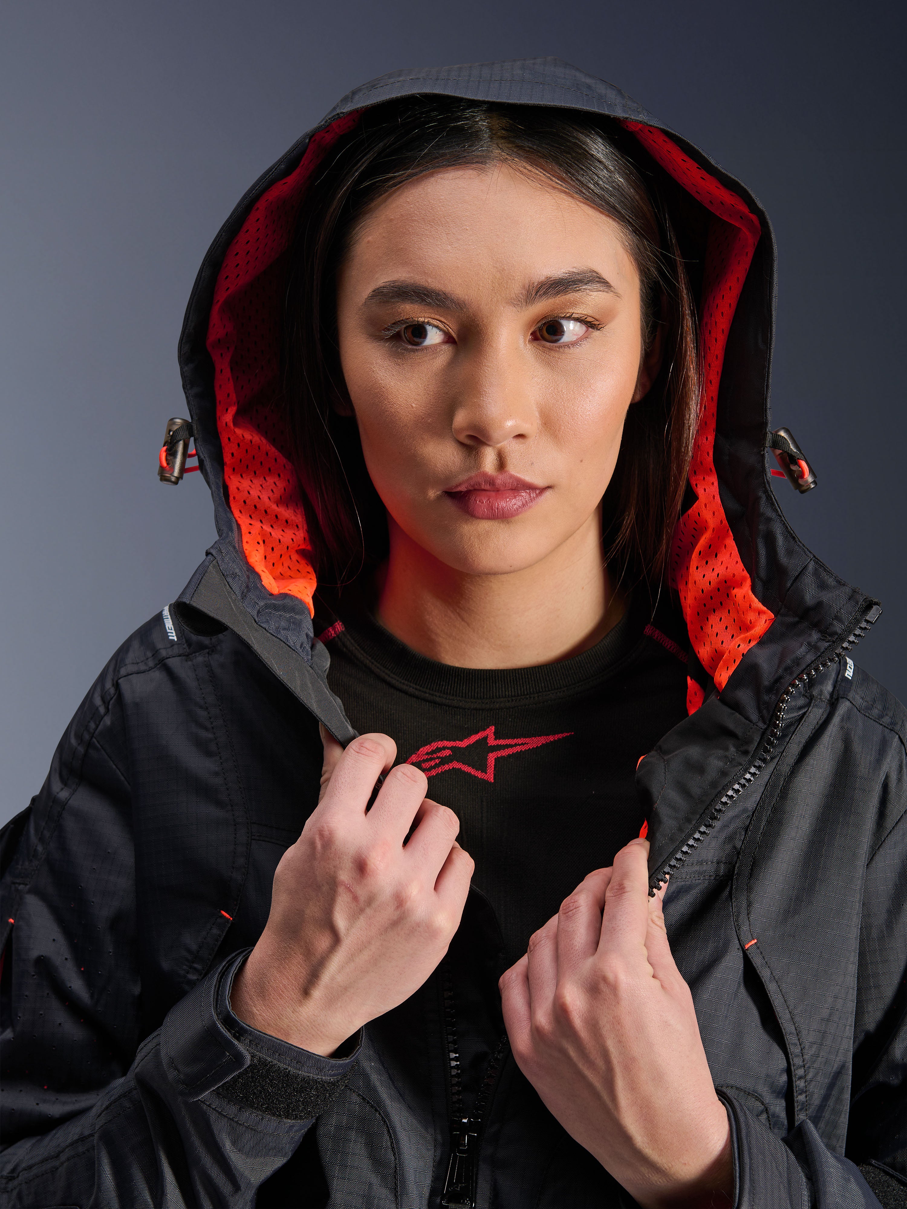 Alpinestars Molly Air Jacket, giacca da moto da donna, nero e corallo fluo, modella con il cappuccio sollevato che mostra la vivace fodera in rete corallo, tessuto tecnico ripstop, abbigliamento da moto urbano contemporaneo
