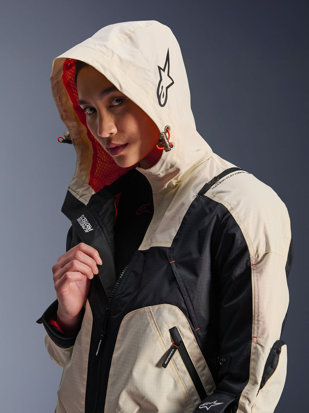 Giacca Alpinestars Molly Air in Black Almond, indossata da una modella con il cappuccio sollevato, caratterizzata da un design color-block crema e nero, fodera in rete arancione vivace ed elementi di design tecnico urbano con logo a stella e dettagli con cerniera.