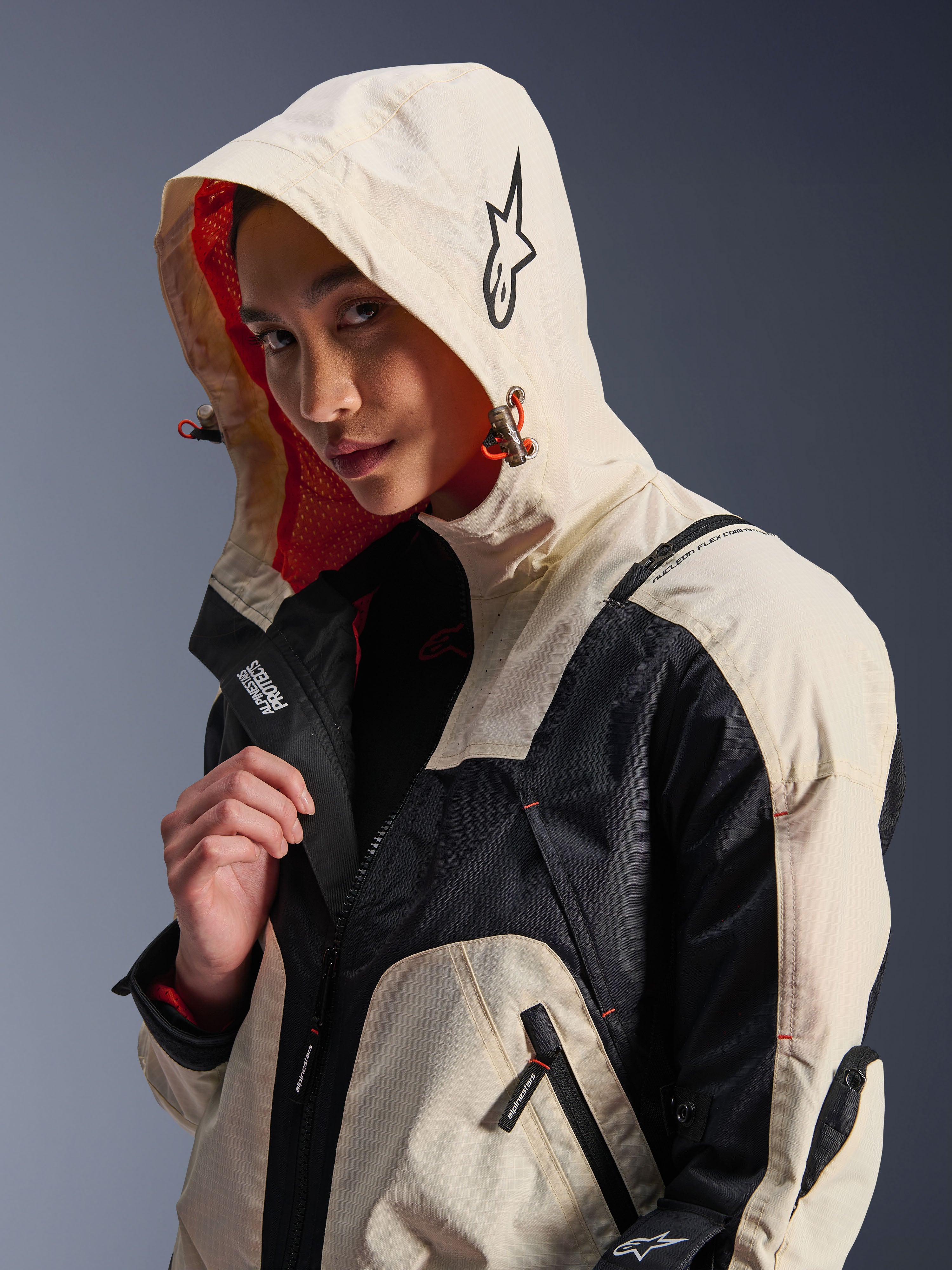 Giacca Alpinestars Molly Air in Black Almond, indossata da una modella con il cappuccio sollevato, caratterizzata da un design color-block crema e nero, fodera in rete arancione vivace ed elementi di design tecnico urbano con logo a stella e dettagli con cerniera.