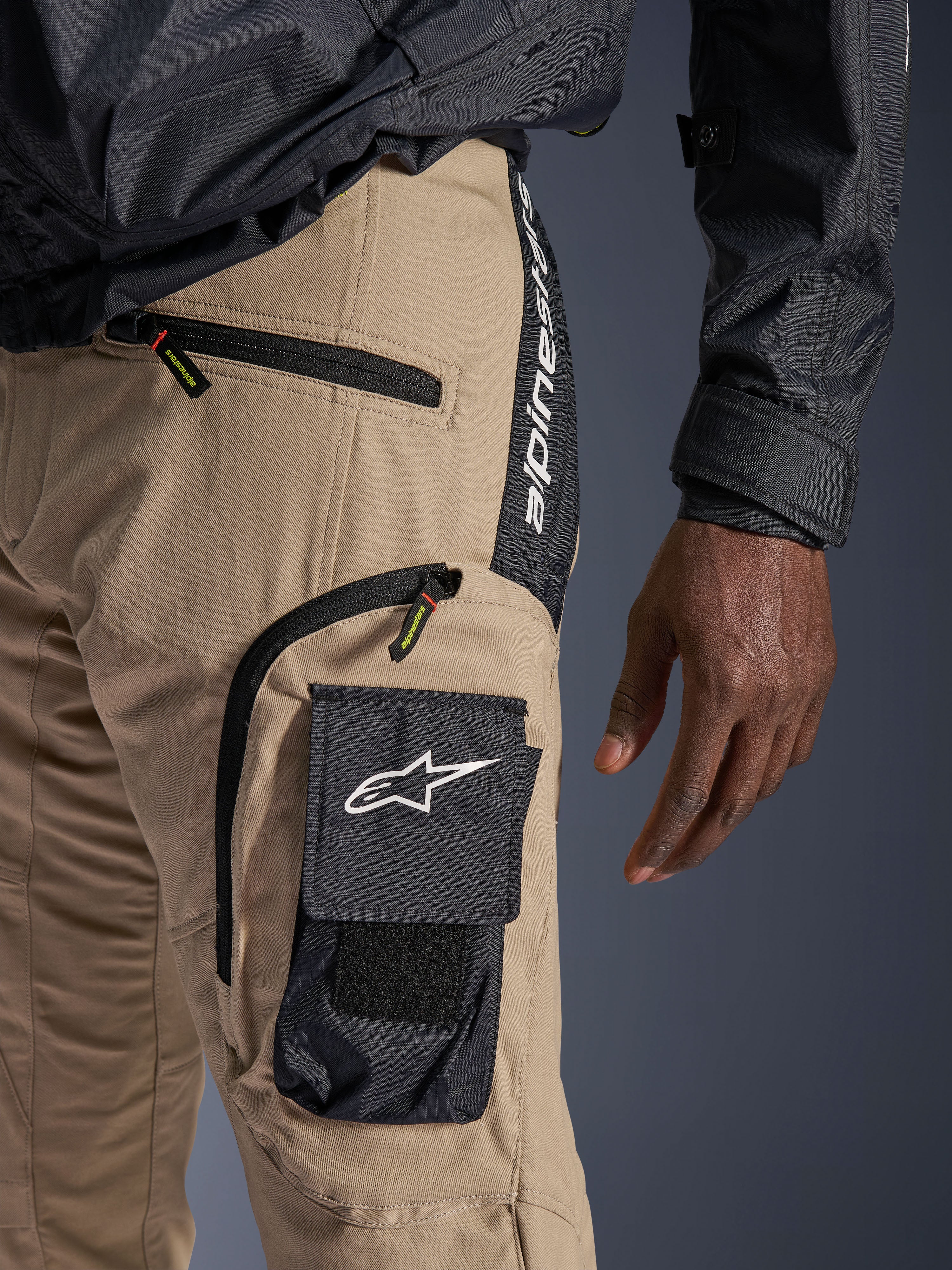 Pantaloni Alpinestars Acteon in colore sabbia, vista dettagliata della zona dell'anca e della coscia, con tasca cargo laterale nera e logo a stella bianco, tasche con cerniera e branding Alpinestars verticale su un pannello laterale nero, abbigliamento da moto urbano.