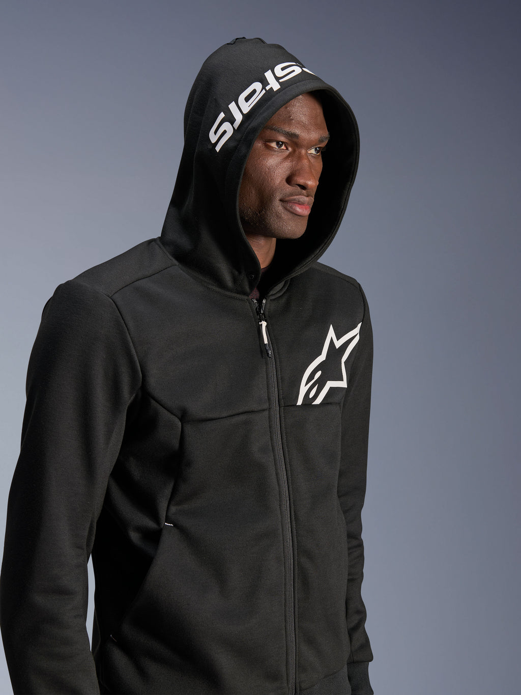 Alpinestars Chrome V2 Sport Hoodie, felpa da moto nera per uso urbano indossata da un modello con cappuccio alzato, logo a stella bianco sul petto e scritta stars bianca sul cappuccio, costruzione in pile tecnico, streetwear casual da moto