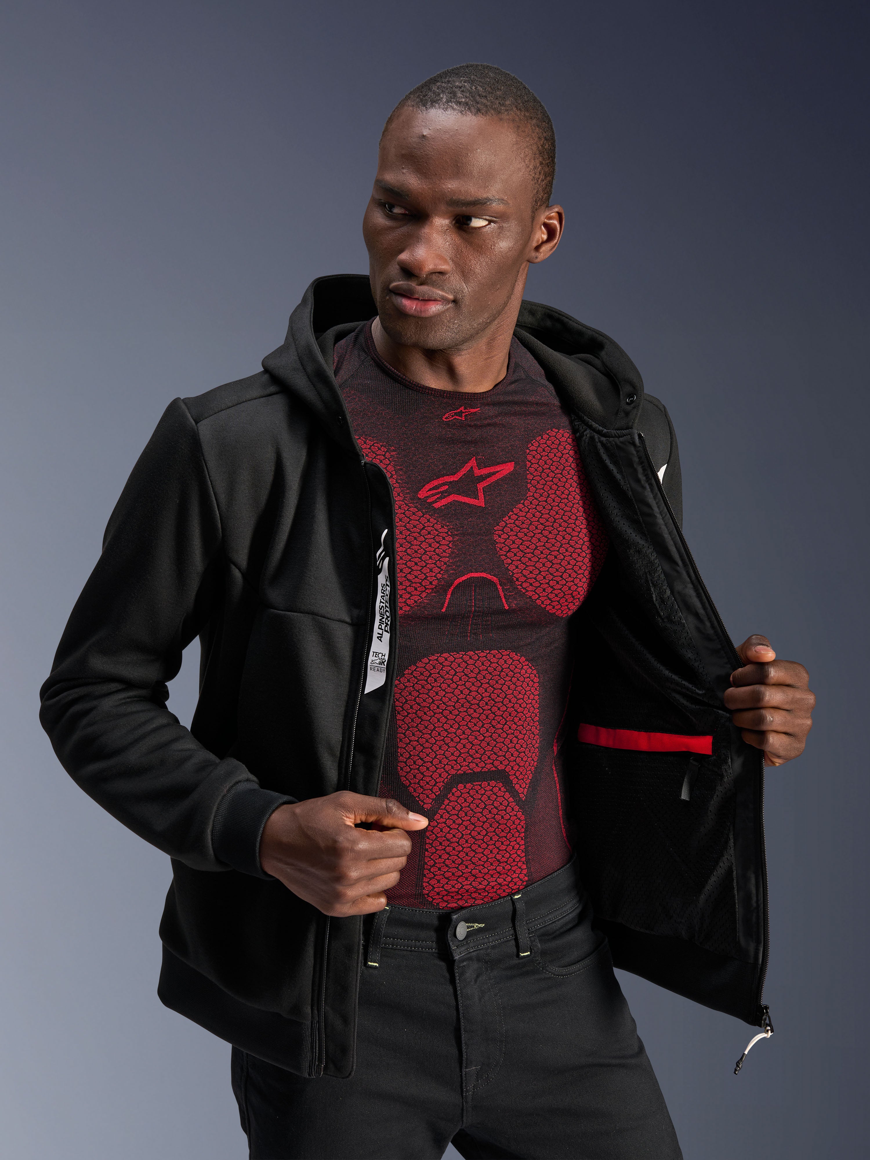 Alpinestars Chrome V2 Sport Hoodie, nero, modello che indossa la giacca aperta per mostrare la fodera interna, la tasca impermeabile rossa e il branding interno, indossata sopra un base layer rosso e nero, abbigliamento da moto urban casual