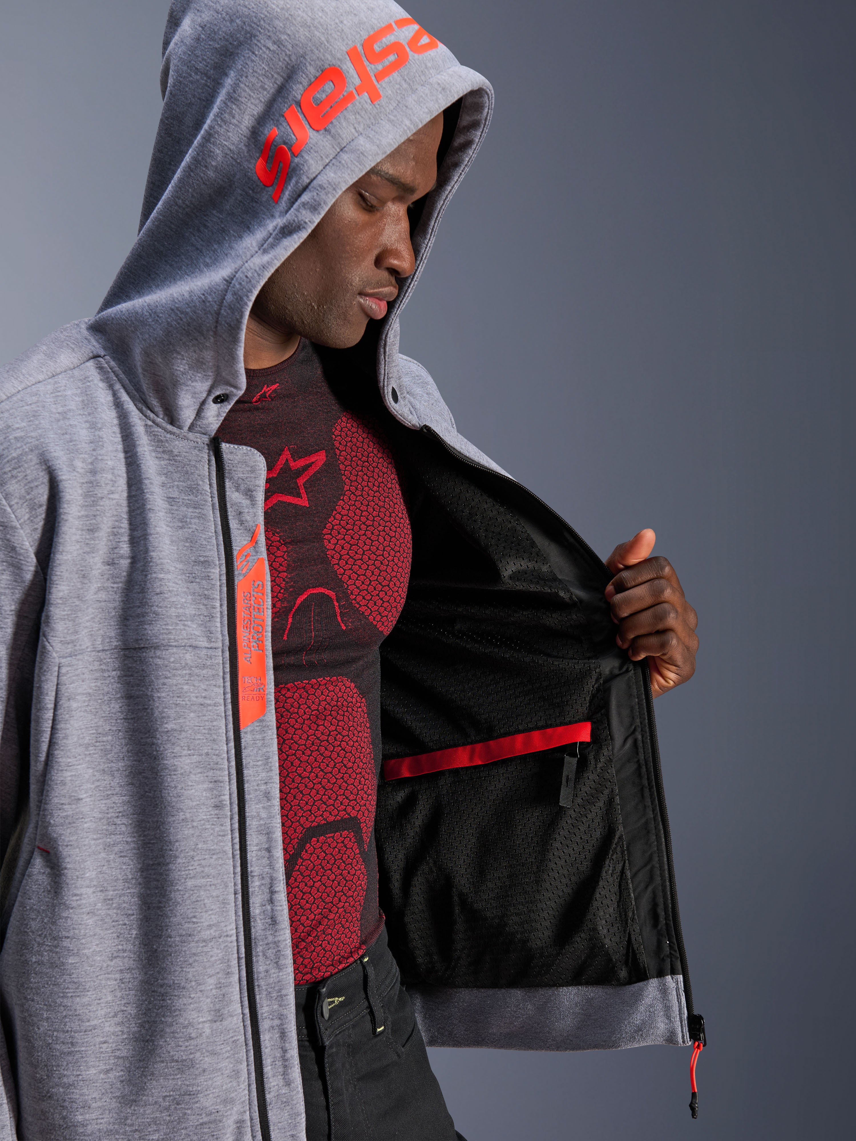 Alpinestars Chrome V2 Sport Hoodie, felpa da moto, grigio mélange e rosso fluo, modello maschile che mostra la fodera interna in rete nera e la tasca impermeabile rossa, caratterizzata da branding rosso sul cappuccio ed etichetta Tech-Air Ready, silhouette atletica casual per la guida urbana