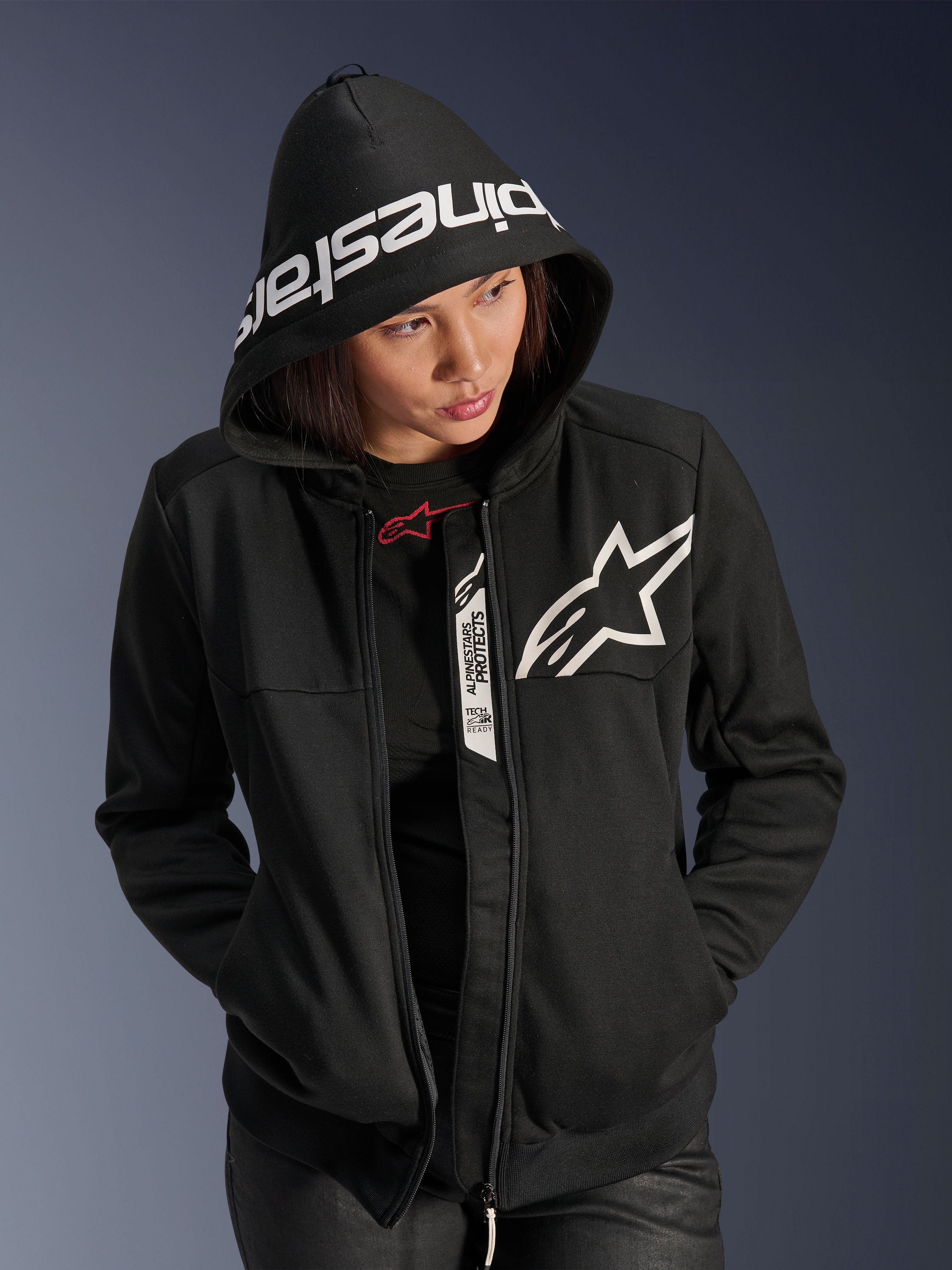 Alpinestars Stella Chrome V2 Sport Hoodie da donna, nero e bianco, felpa da moto con cappuccio, logo a stella bianco sul petto e scritta Alpinestars sul bordo del cappuccio, modella con cappuccio alzato, stile sportivo urbano casual con dettagli Tech-Air Ready