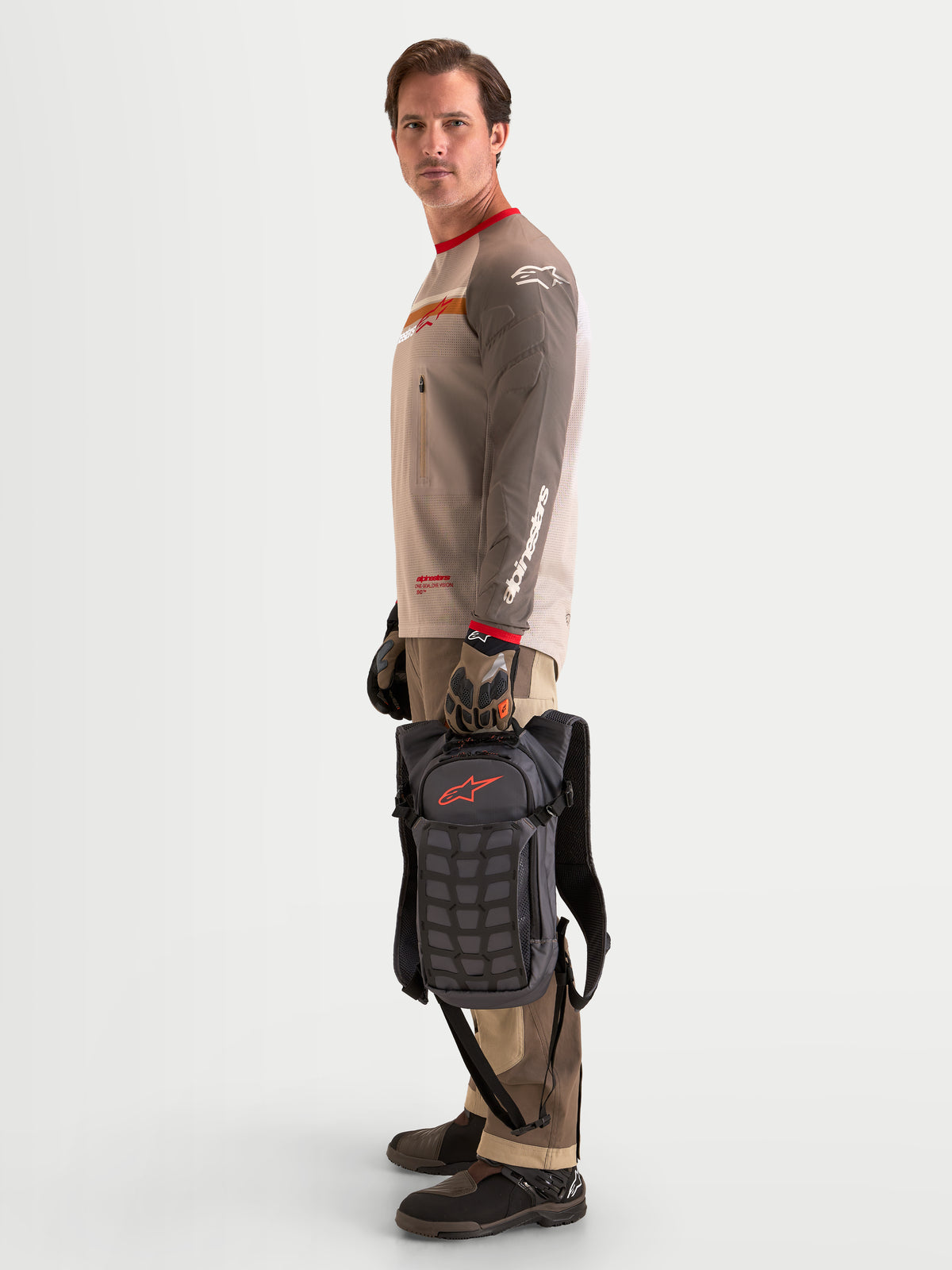 Alpinestars Zaino AT-9, zaino da motocross in marrone noce e grigio scuro, vista laterale di un modello in piedi che tiene la borsa per il manico superiore, mostrando i pannelli strutturati e gli scomparti organizzati