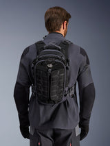 Alpinestars AT-15 Back Pack in Black Deep Gray, zaino tecnico da avventura indossato da un pilota visto di schiena, con pannelli frontali testurizzati e logo 'ALL TERRA', abbinato a una giacca da moto grigia e nera.