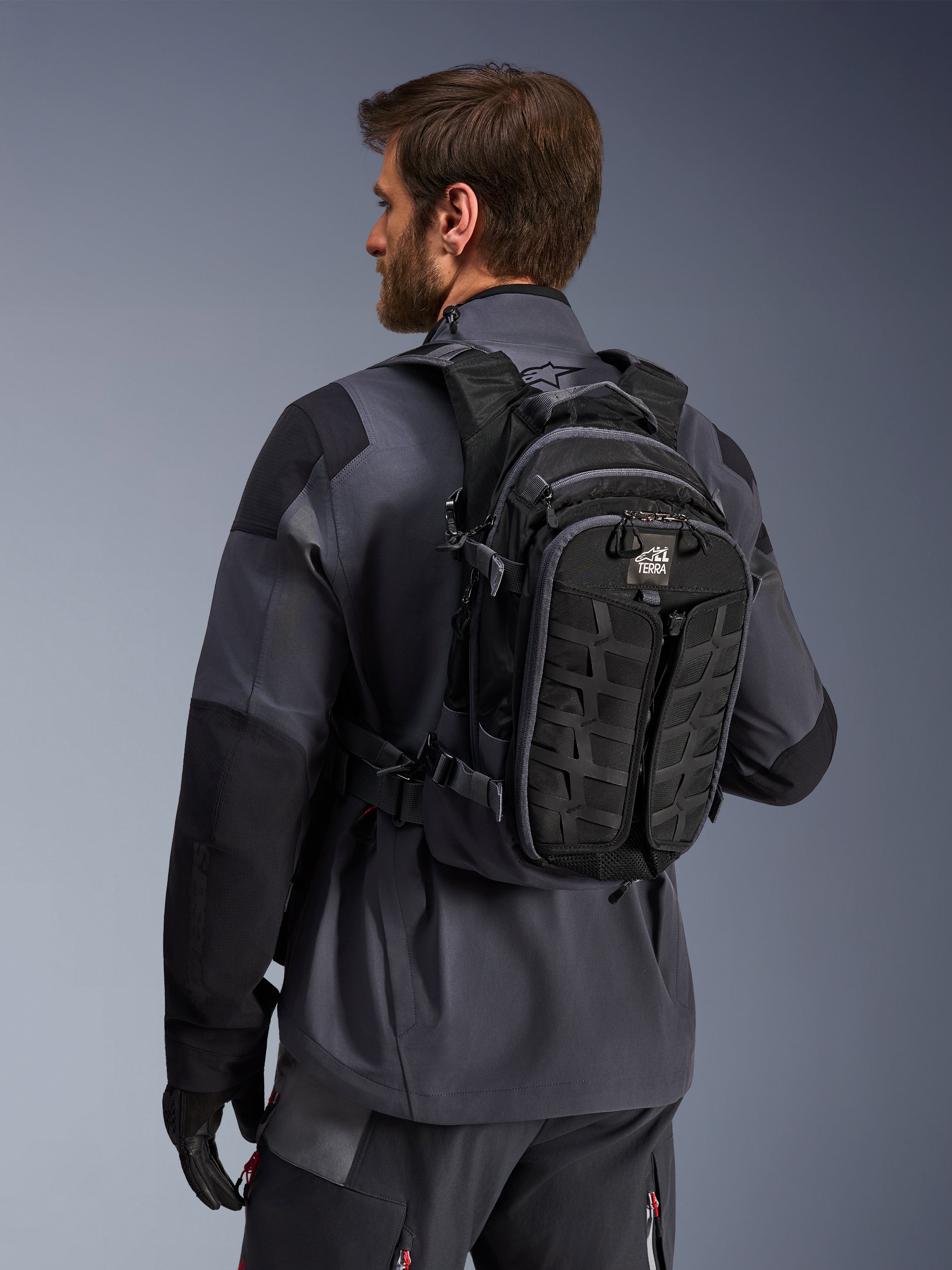Alpinestars AT-15 Back Pack, zaino da moto adventure, nero e grigio scuro, indossato da un pilota con giacca grigia, caratterizzato da un design tecnico con sistema MOLLE e marchio 'TERRA', equipaggiamento ergonomico per il fuoristrada