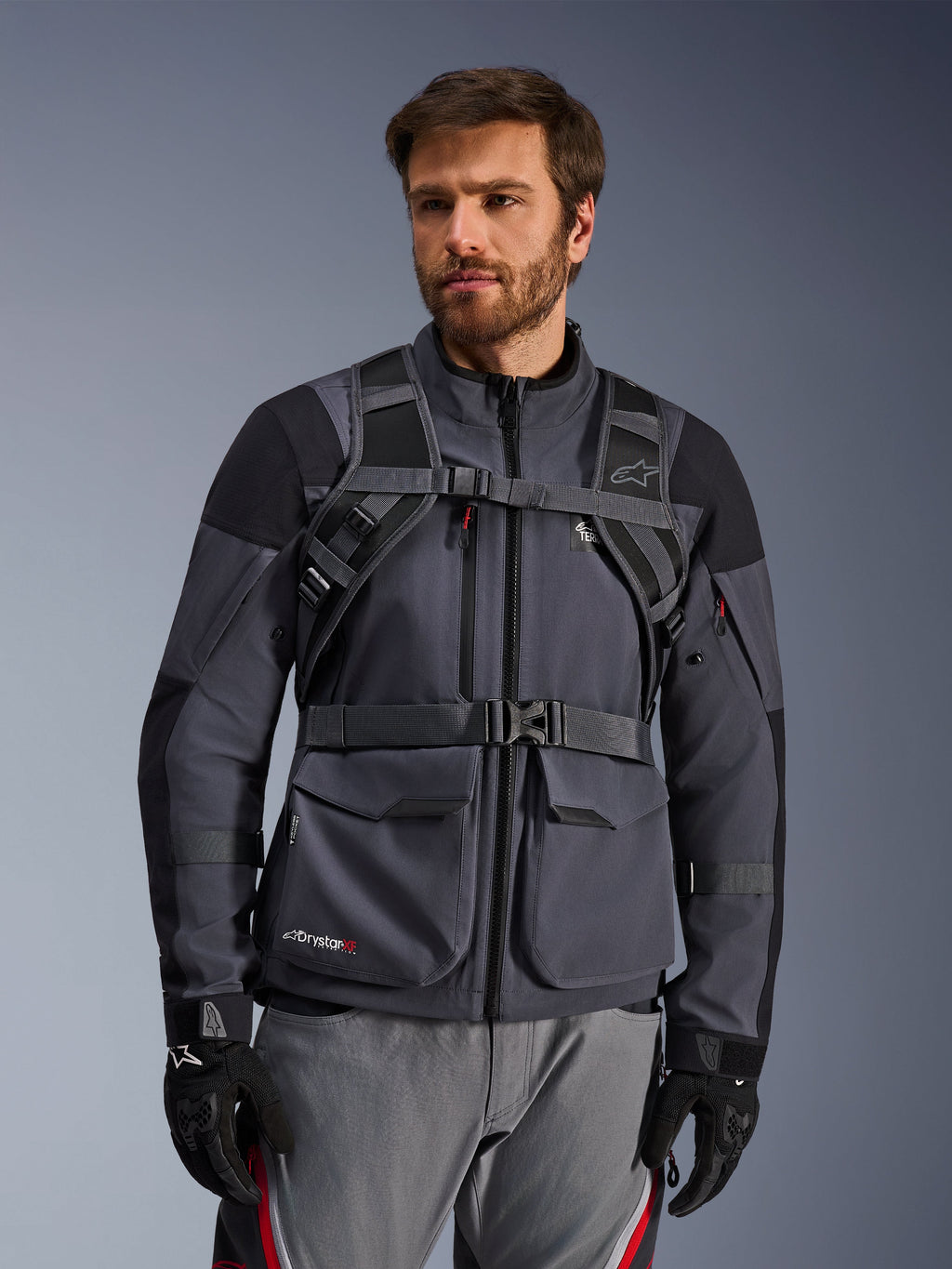 Sistema di imbracatura dello zaino Alpinestars AT-15, Black Deep Gray, vista frontale indossato da un pilota sopra una giacca da moto grigia, con chiusura magnetica sul petto e cinghia in vita sicura, abbinato a guanti neri per il motociclismo adventure