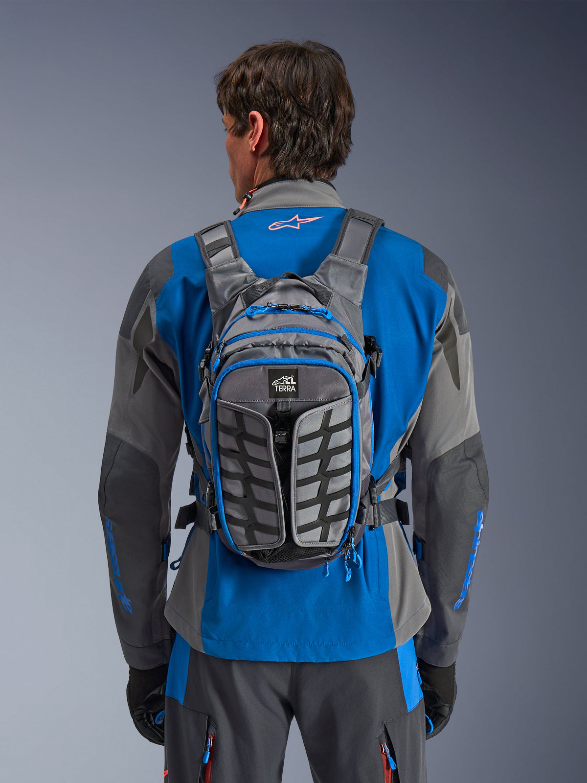 Alpinestars AT-15 Back Pack, zaino da moto adventure, grigio scuro e grigio intenso, indossato da un pilota sopra una giacca blu e grigia, con marchio All Terra, design strutturale ergonomico, cinghie regolabili e dettagli delle cerniere blu.