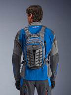 Alpinestars AT-15 Back Pack, zaino da moto adventure, grigio scuro e grigio intenso, indossato da un pilota sopra una giacca blu e grigia, con marchio All Terra, design strutturale ergonomico, cinghie regolabili e dettagli delle cerniere blu.