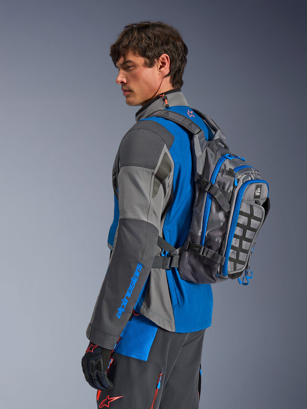 Alpinestars AT-15 Back Pack, zaino da moto adventure, grigio scuro con accenti blu, indossato da un pilota con abbigliamento tecnico coordinato grigio e blu, caratterizzato da pannello MOLLE e struttura robusta per avventure off-road.