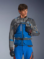 Zaino Alpinestars AT-15, zaino da moto, grigio scuro, indossato da un pilota con giacca e pantaloni adventure blu e grigi, con spallacci tecnici grigi, fibbia magnetica sul petto e cinghia in vita, design robusto per la guida off-road