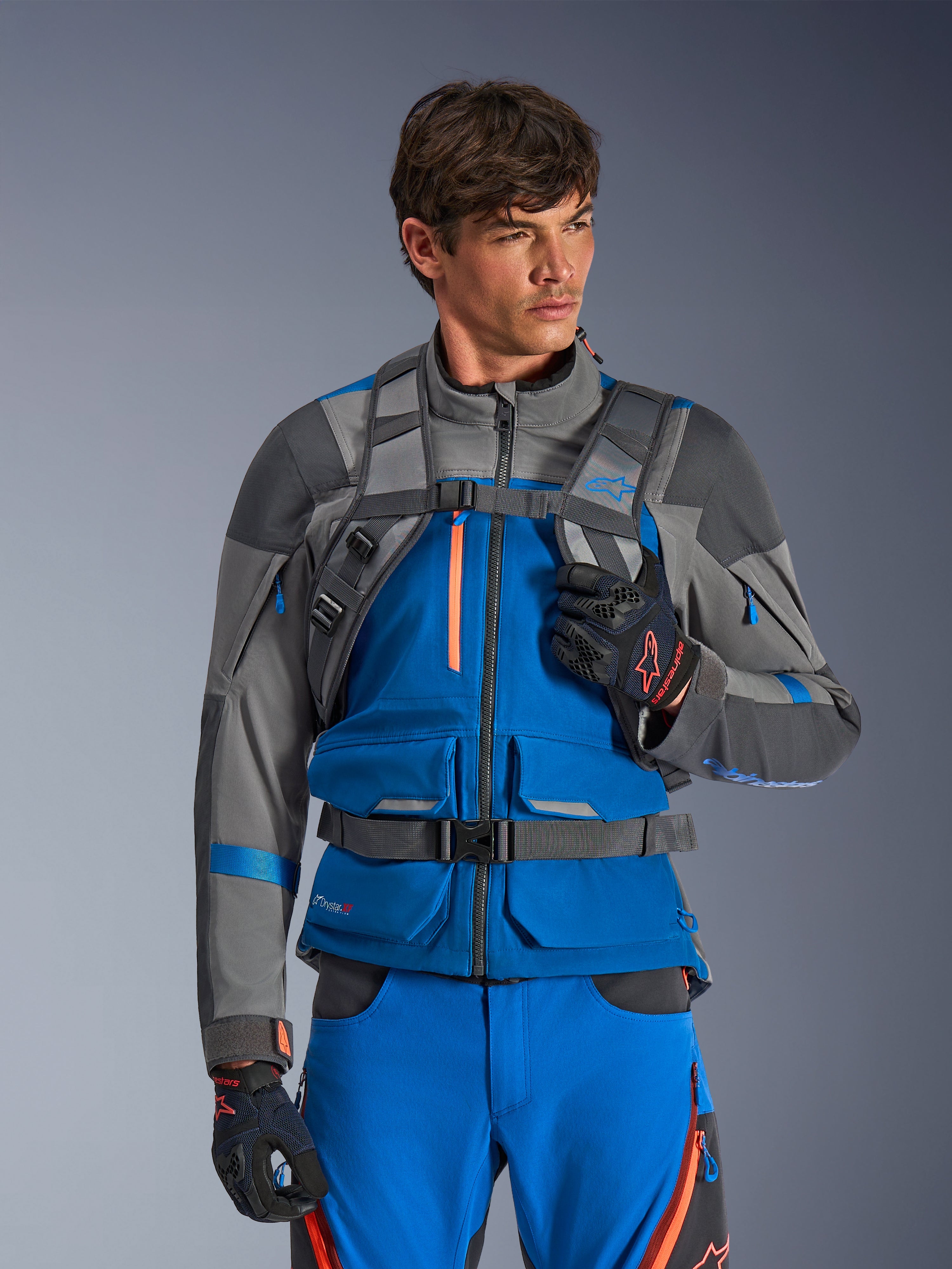 Zaino Alpinestars AT-15, zaino da moto, grigio scuro, indossato da un pilota con giacca e pantaloni adventure blu e grigi, con spallacci tecnici grigi, fibbia magnetica sul petto e cinghia in vita, design robusto per la guida off-road