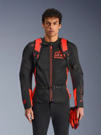 Uomo che indossa lo zaino Alpinestars MM93 AMP3, rosso fluo e nero, con spallacci regolabili e cinturino pettorale con fibbia, abbinato a giacca da moto e guanti MM93 coordinati, equipaggiamento tecnico della collezione Marc Marquez