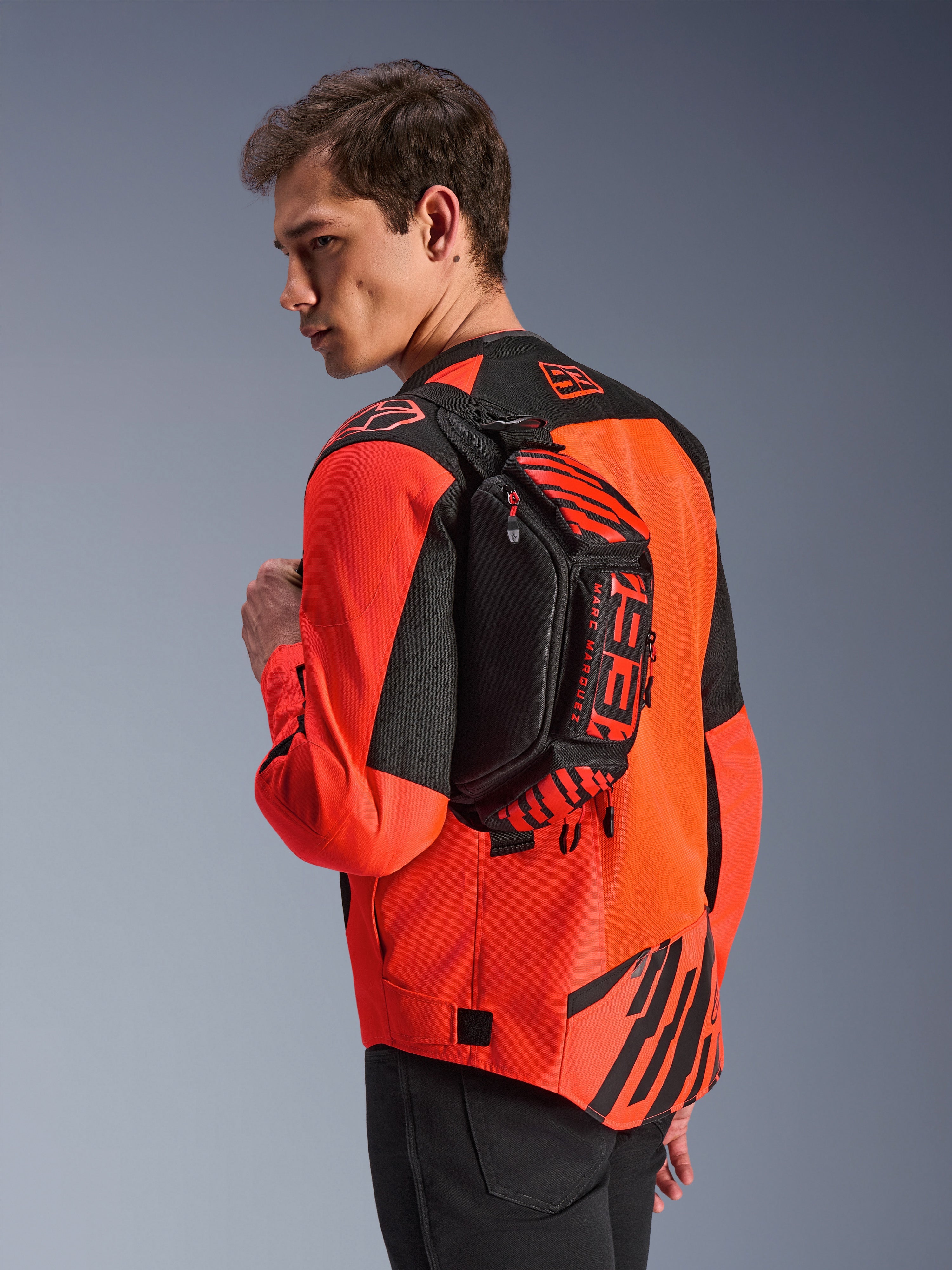 Alpinestars MM93 Waist Bag, marsupio nero con dettagli rosso fluo e rosso brillante, indossato da un modello sulla schiena sopra una giacca da moto rossa e nera, con loghi MM93 e grafiche Marc Marquez