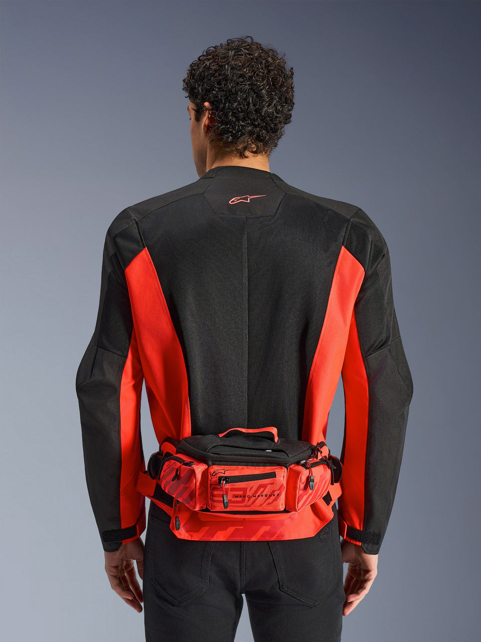 Alpinestars MM93 Waist Bag, rosso e nero, indossato sopra una giacca da moto nera e rossa, con marchio Marc Marquez 93, cintura regolabile e scomparti con zip, accessorio funzionale d'ispirazione racing