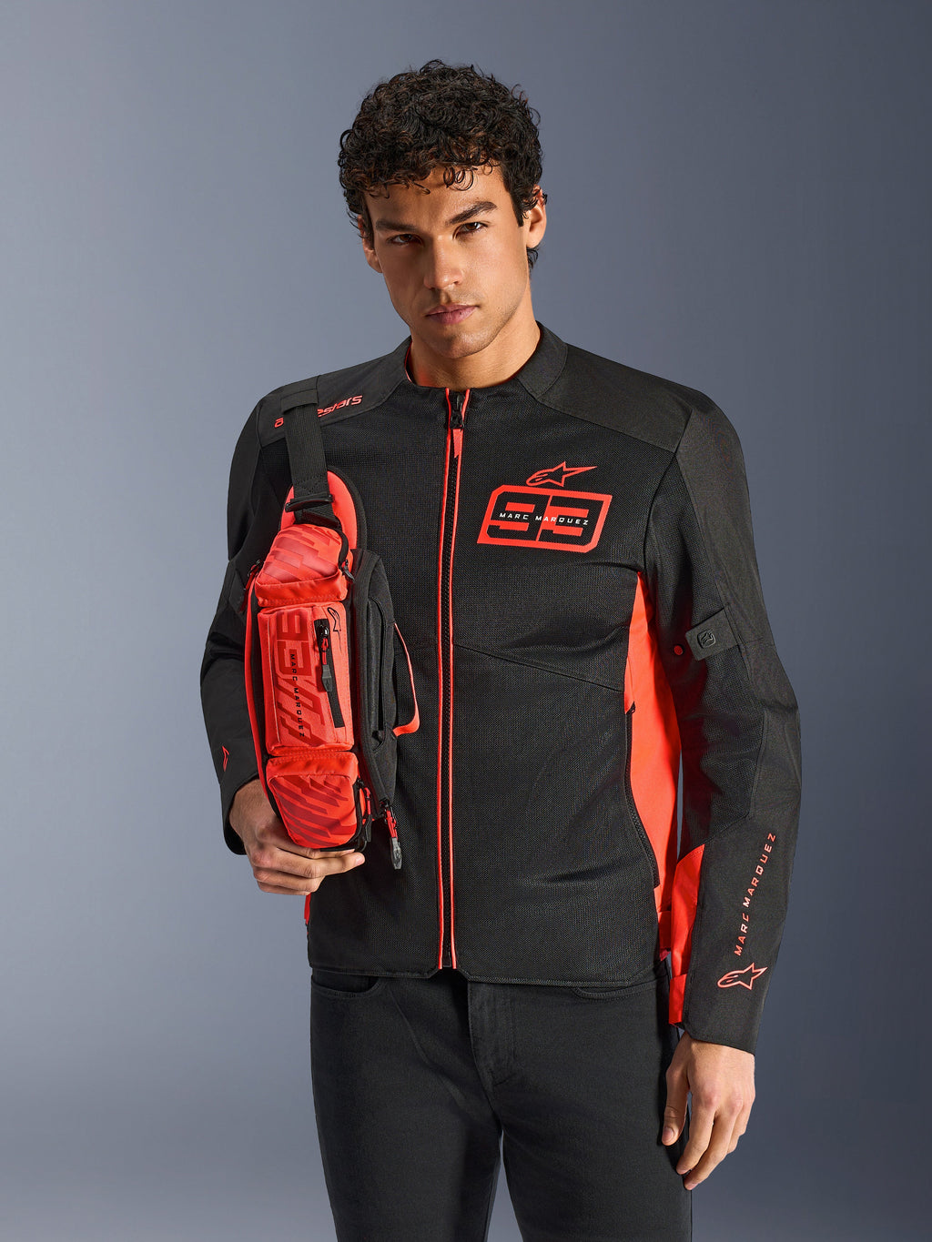Uomo con borsa a vita Alpinestars MM93, rosso fluo e nero, indossata a tracolla sopra una giacca da moto coordinata nera e rossa, con logo Marc Marquez 93, design sportivo racing
