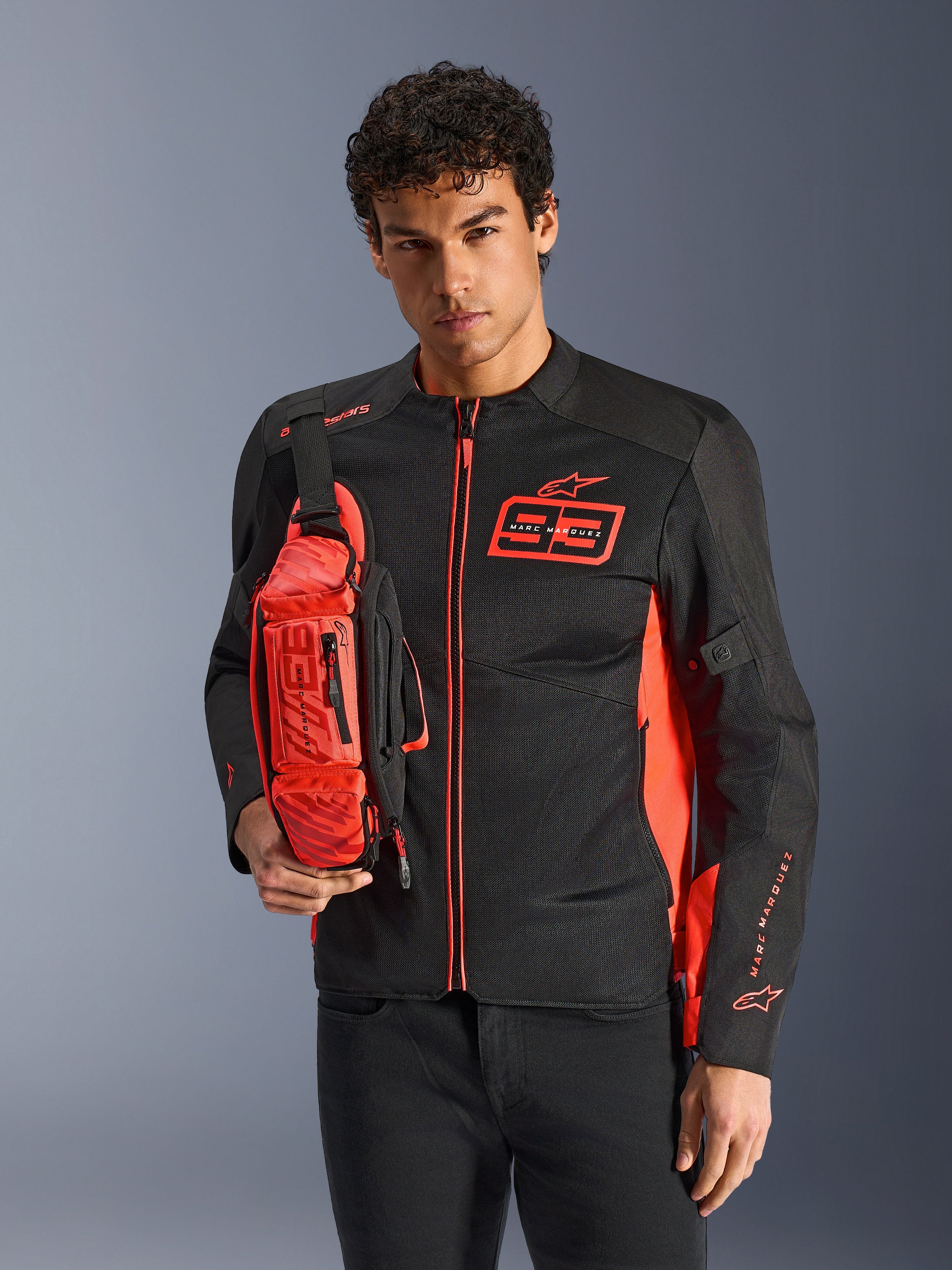 Uomo con borsa a vita Alpinestars MM93, rosso fluo e nero, indossata a tracolla sopra una giacca da moto coordinata nera e rossa, con logo Marc Marquez 93, design sportivo racing