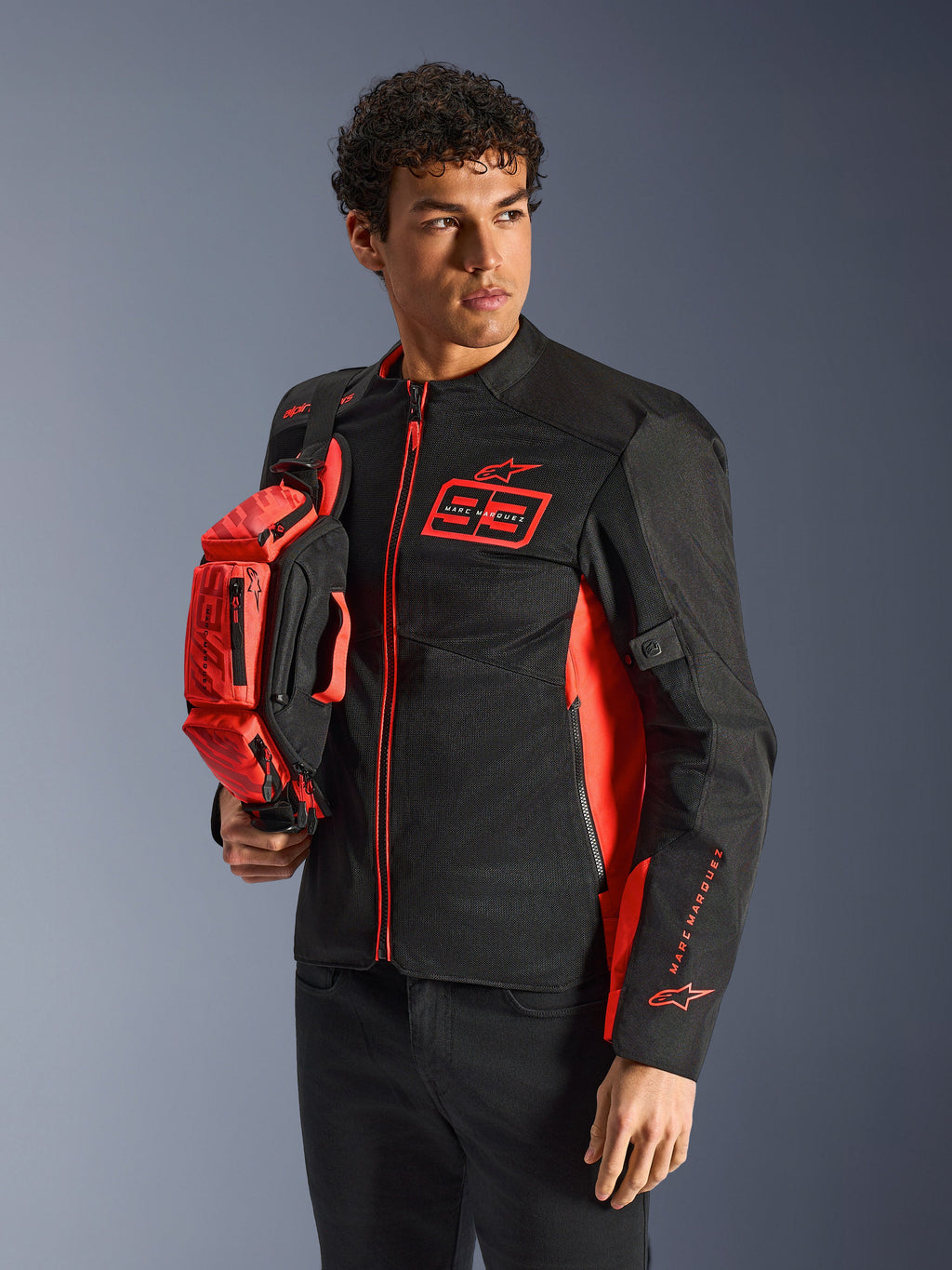 Alpinestars MM93 Waist Bag, borsa a vita in rosso fluo e nero, tenuta da un modello con giacca da moto coordinata nera e rossa, dotata di più scomparti con zip e logo Marc Marquez, accessorio pratico per la guida sportiva