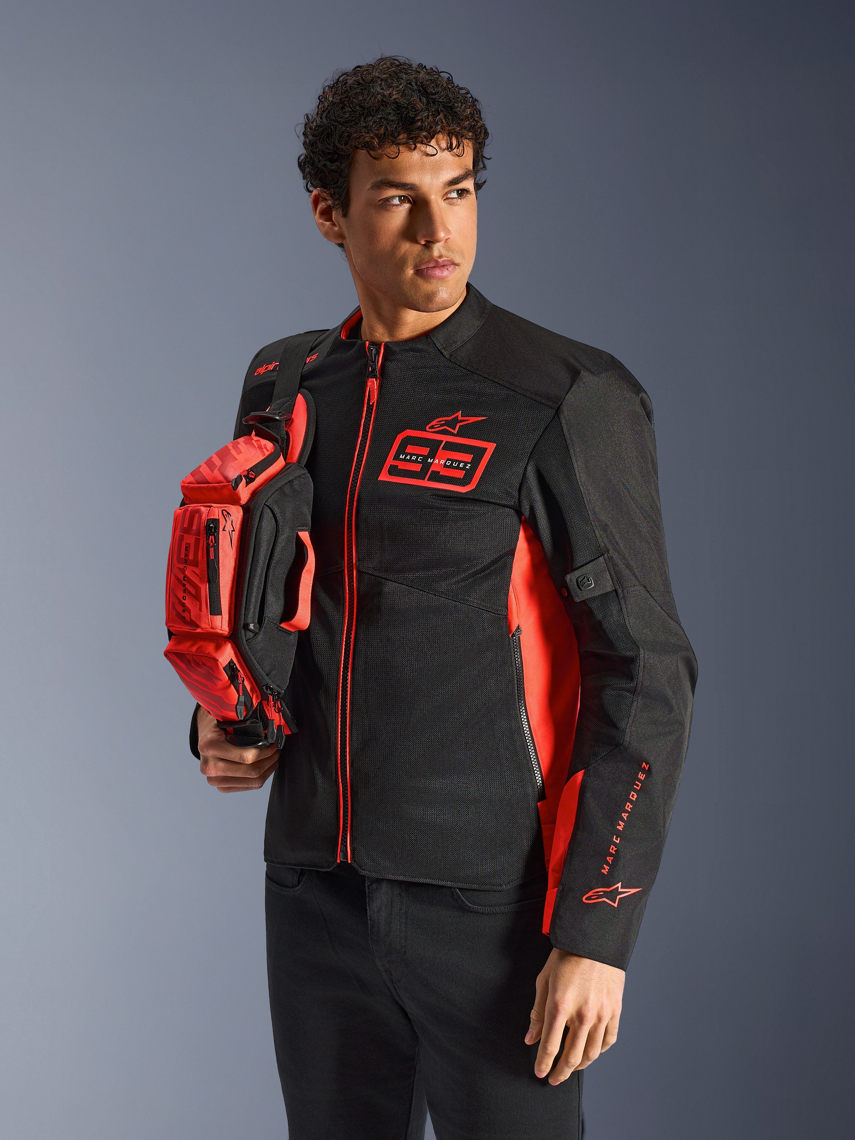 Alpinestars MM93 Waist Bag, borsa a vita in rosso fluo e nero, tenuta da un modello con giacca da moto coordinata nera e rossa, dotata di più scomparti con zip e logo Marc Marquez, accessorio pratico per la guida sportiva