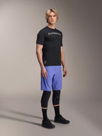 Modello che indossa abbigliamento da mountain bike Alpinestars, con la maglia a maniche corte A-Aria Switch nera e i pantaloncini A-Aria Lite Baja Blue, abbinati a ginocchiere nere, calze nere e scarpe da mountain bike nere in uno scatto in studio a figura intera.