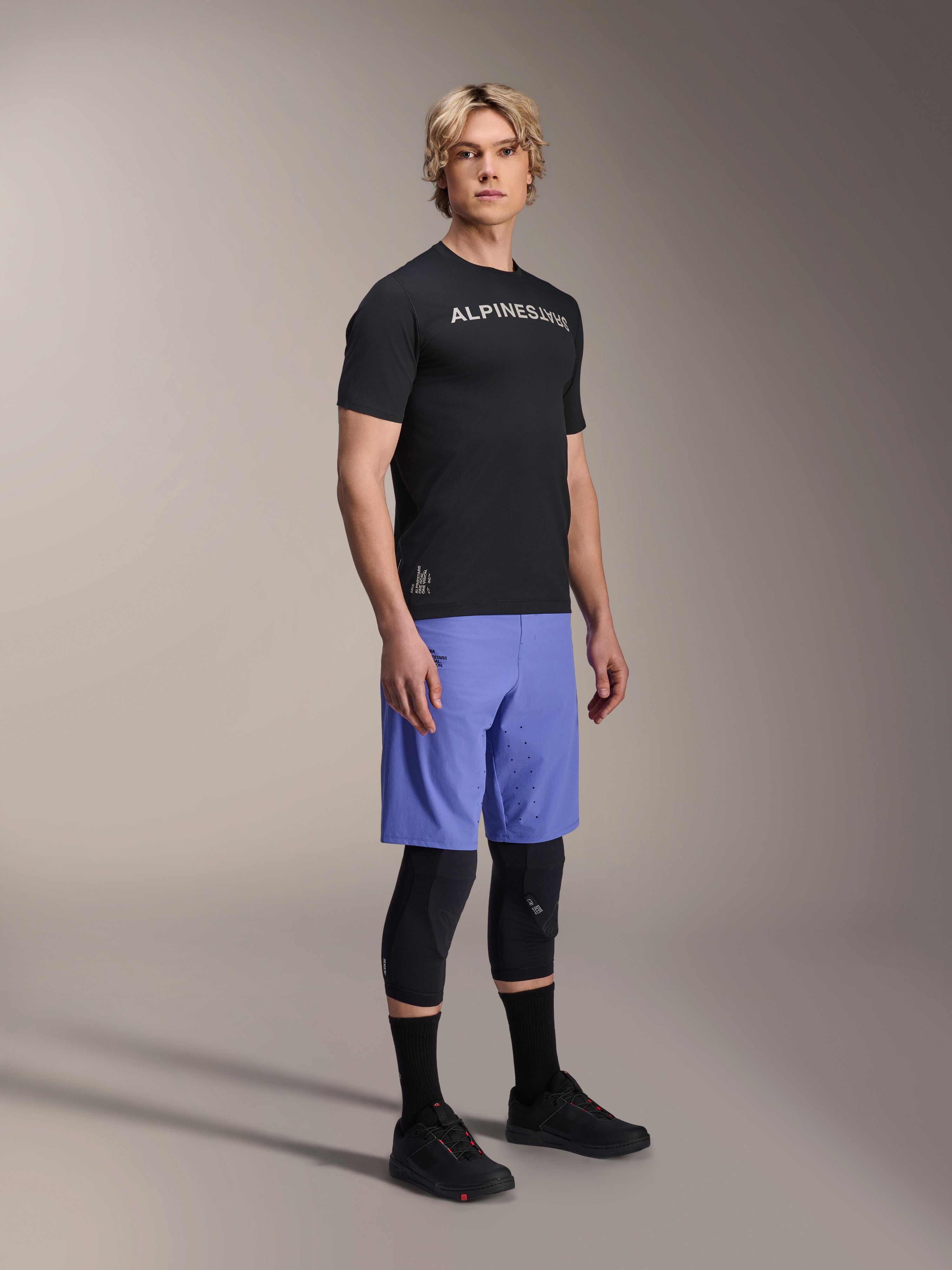 Modello che indossa abbigliamento da mountain bike Alpinestars, con la maglia a maniche corte A-Aria Switch nera e i pantaloncini A-Aria Lite Baja Blue, abbinati a ginocchiere nere, calze nere e scarpe da mountain bike nere in uno scatto in studio a figura intera.