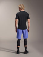 Rider uomo che indossa i pantaloncini Alpinestars A-Aria Lite, pantaloncini da MTB, Baja Blue, vista posteriore, abbinati a una maglia nera a maniche corte, ginocchiere nere e scarpe da ciclismo nere, abbigliamento per mountain bike ad alte prestazioni