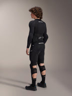 Modello che indossa protezioni ginocchio e stinco Alpinestars A-Impact Plasma Pro, vista dal retro, nero e bianco, con chiusura a triplo cinturino e pannelli in rete ventilata, indossate con un set completo di protezioni da mountain bike nero, base layer a maniche lunghe e pantaloncini imbottiti
