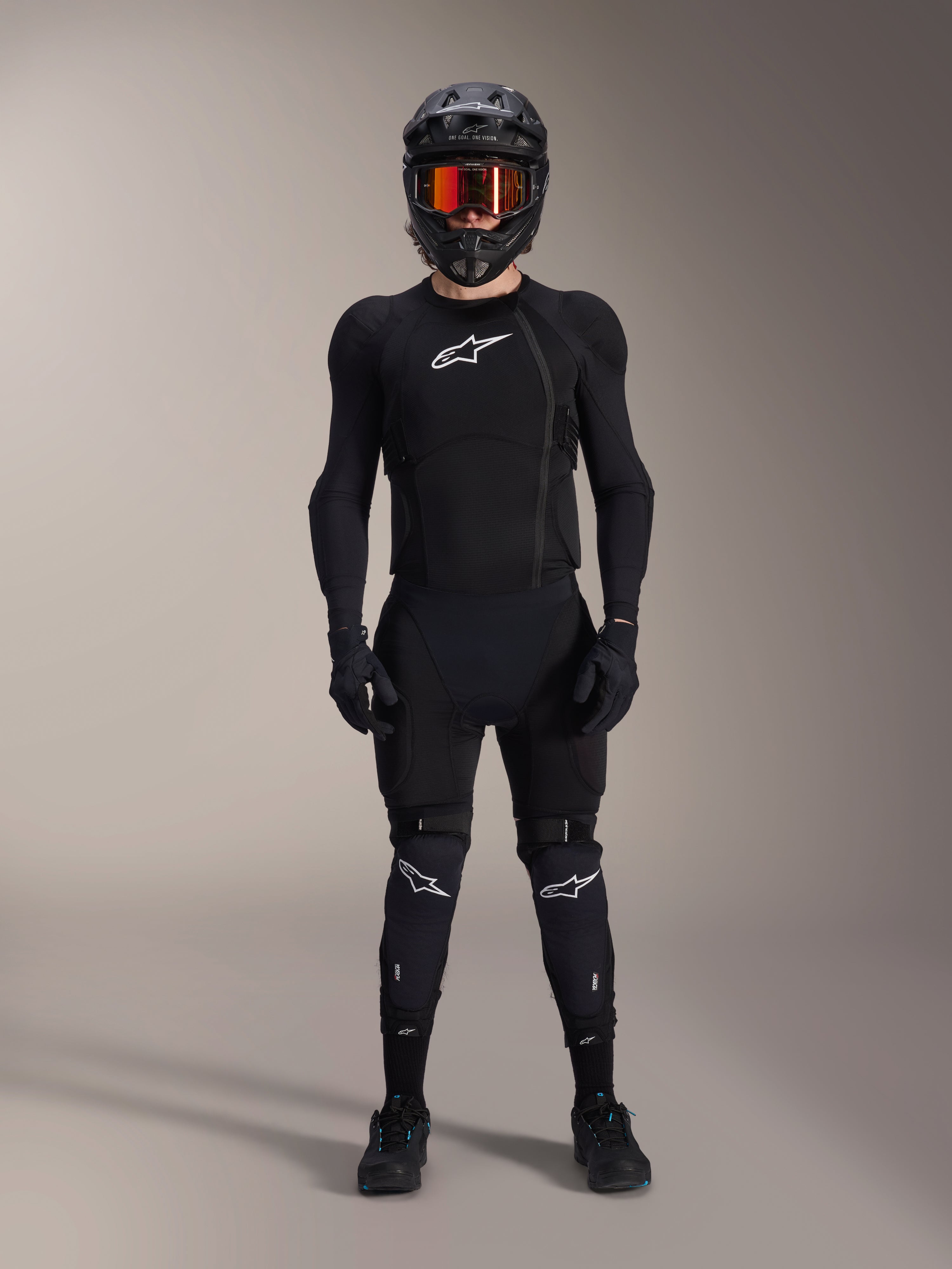 Ciclista di mountain bike con equipaggiamento protettivo completo, Giacca protettiva Alpinestars A-Impact Plasma in Nero e Bianco, con logo bianco sul petto e tessuto mesh, indossata con casco nero, maschera con lenti rosse e ginocchiere, set coordinato di protezioni MTB ad alte prestazioni.