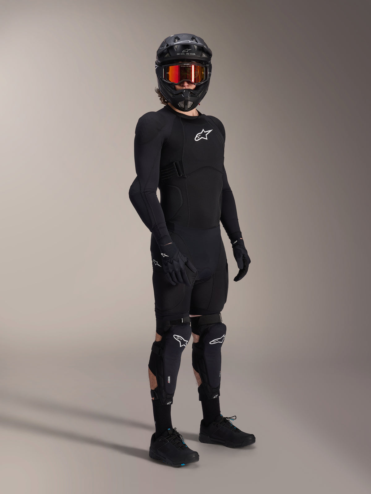 Protezioni ginocchio e tibia Alpinestars A-Impact Plasma Pro, nero e bianco, indossate da un rider con abbigliamento protettivo completo da mountain bike, maglia protettiva nera a maniche lunghe, guanti neri e casco integrale con maschera arancione