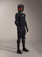 Protezioni ginocchio e tibia Alpinestars A-Impact Plasma Pro, nero e bianco, indossate da un rider con abbigliamento protettivo completo da mountain bike, maglia protettiva nera a maniche lunghe, guanti neri e casco integrale con maschera arancione