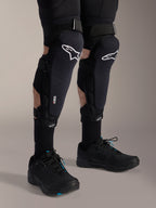 Alpinestars A-Impact Plasma Pro Knee Shin Protectors, nero e bianco, con logo Alpinestars bianco e sistema di chiusura a triplo cinturino, indossati con calze nere e scarpe sportive nere, protezione durevole per il ciclismo fuoristrada