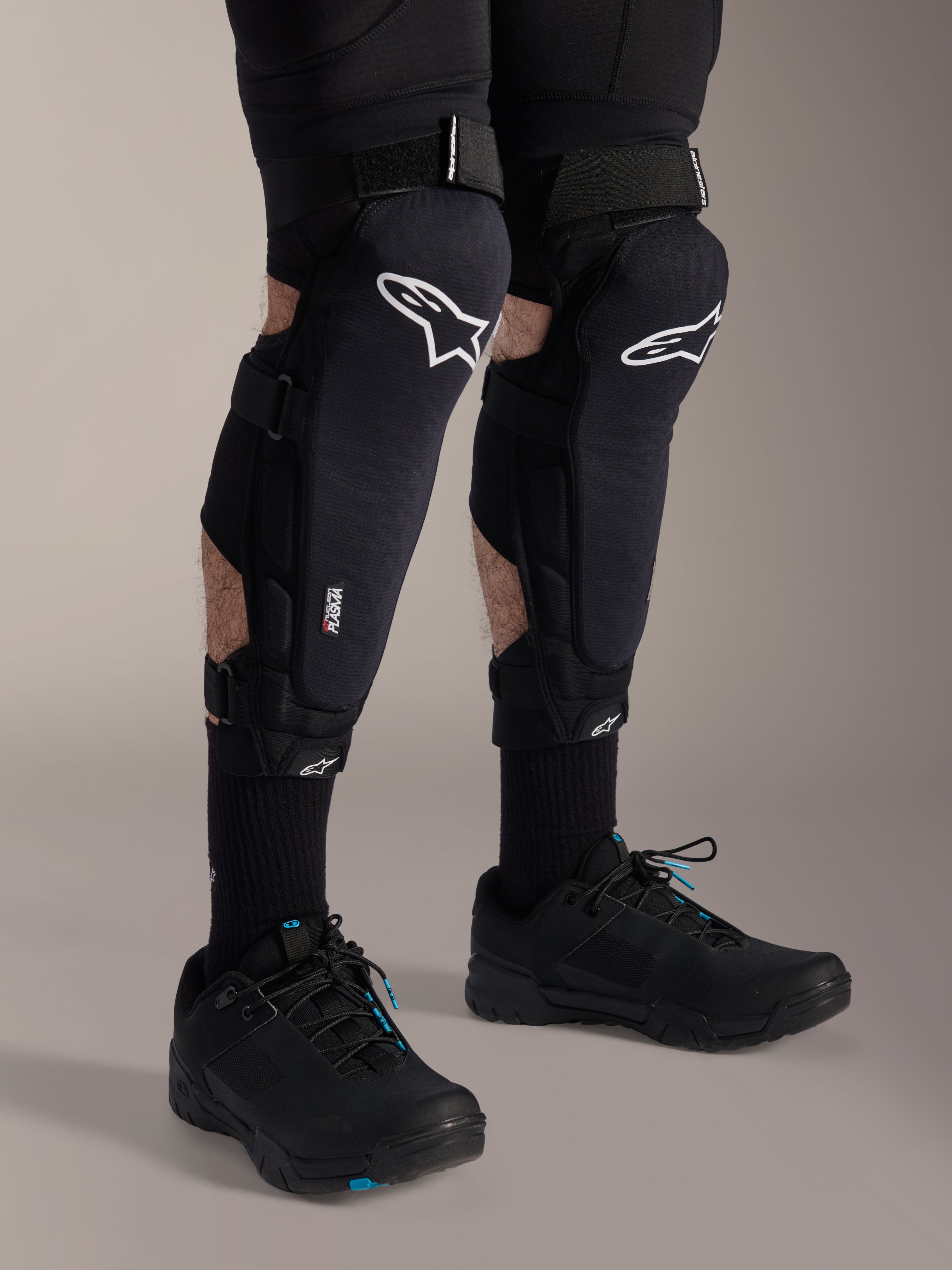 Ginocchiere Alpinestars A-Impact Plasma Pro, nero e bianco, con logo della tecnologia Nucleon PLASMA, indossate con calze nere e scarpe da mountain bike nere, chiusure a strappo sicure, equipaggiamento protettivo ergonomico per ciclismo fuoristrada