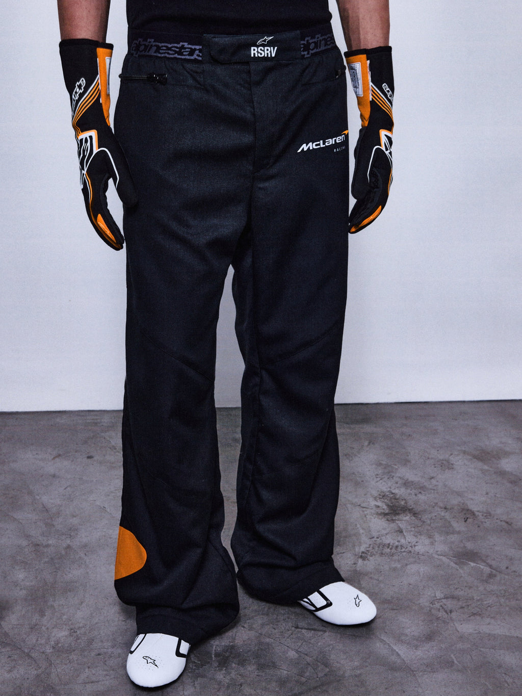 Alpinestars RSRV Arena Suit Pants, pantaloni da corsa neri con loghi McLaren e Alpinestars, dettagli arancioni, indossati con guanti racing neri e arancioni e scarpe bianche con accenti neri