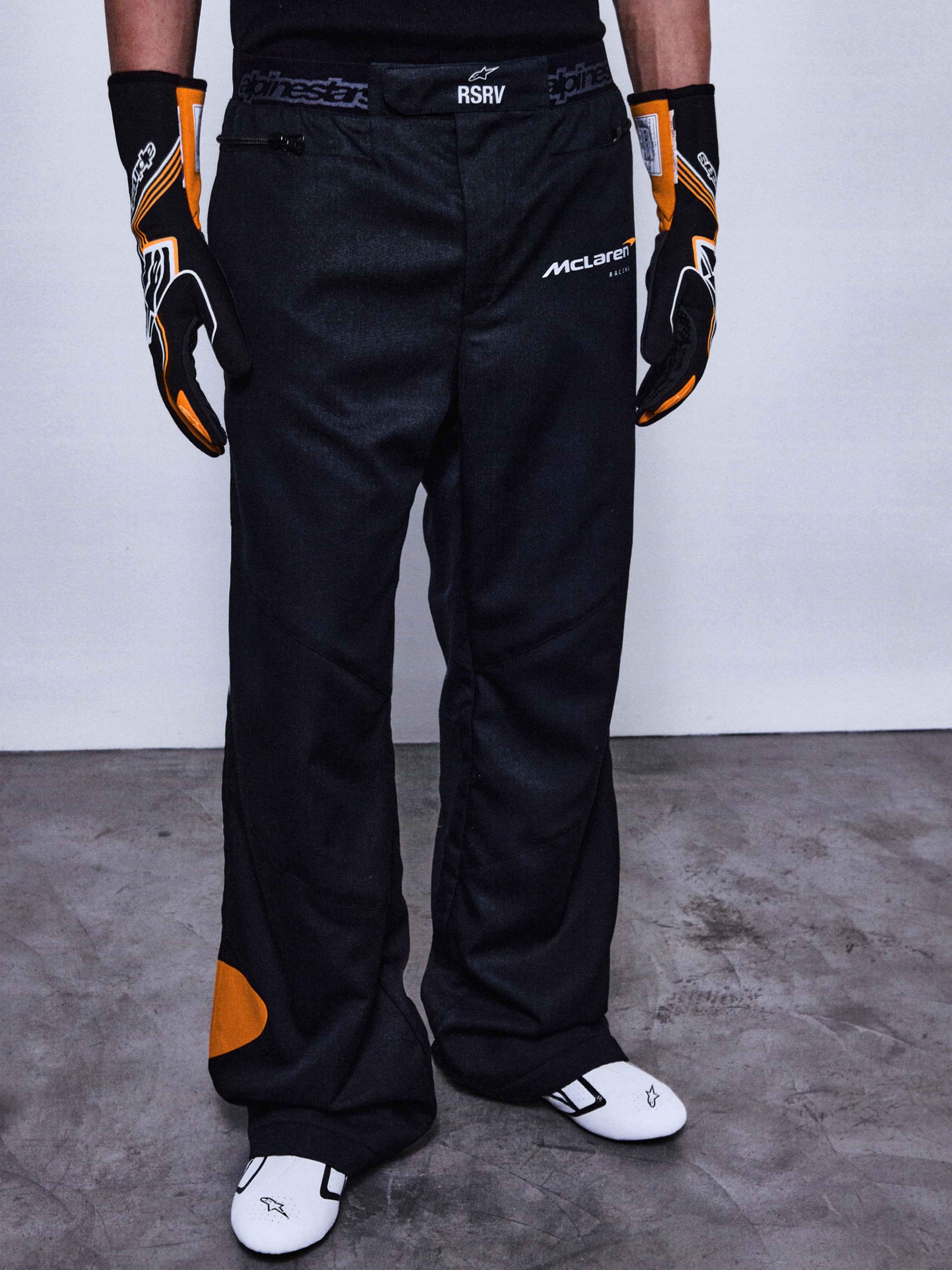 Alpinestars RSRV Arena Suit Pants, pantaloni da corsa neri con loghi McLaren e Alpinestars, dettagli arancioni, indossati con guanti racing neri e arancioni e scarpe bianche con accenti neri