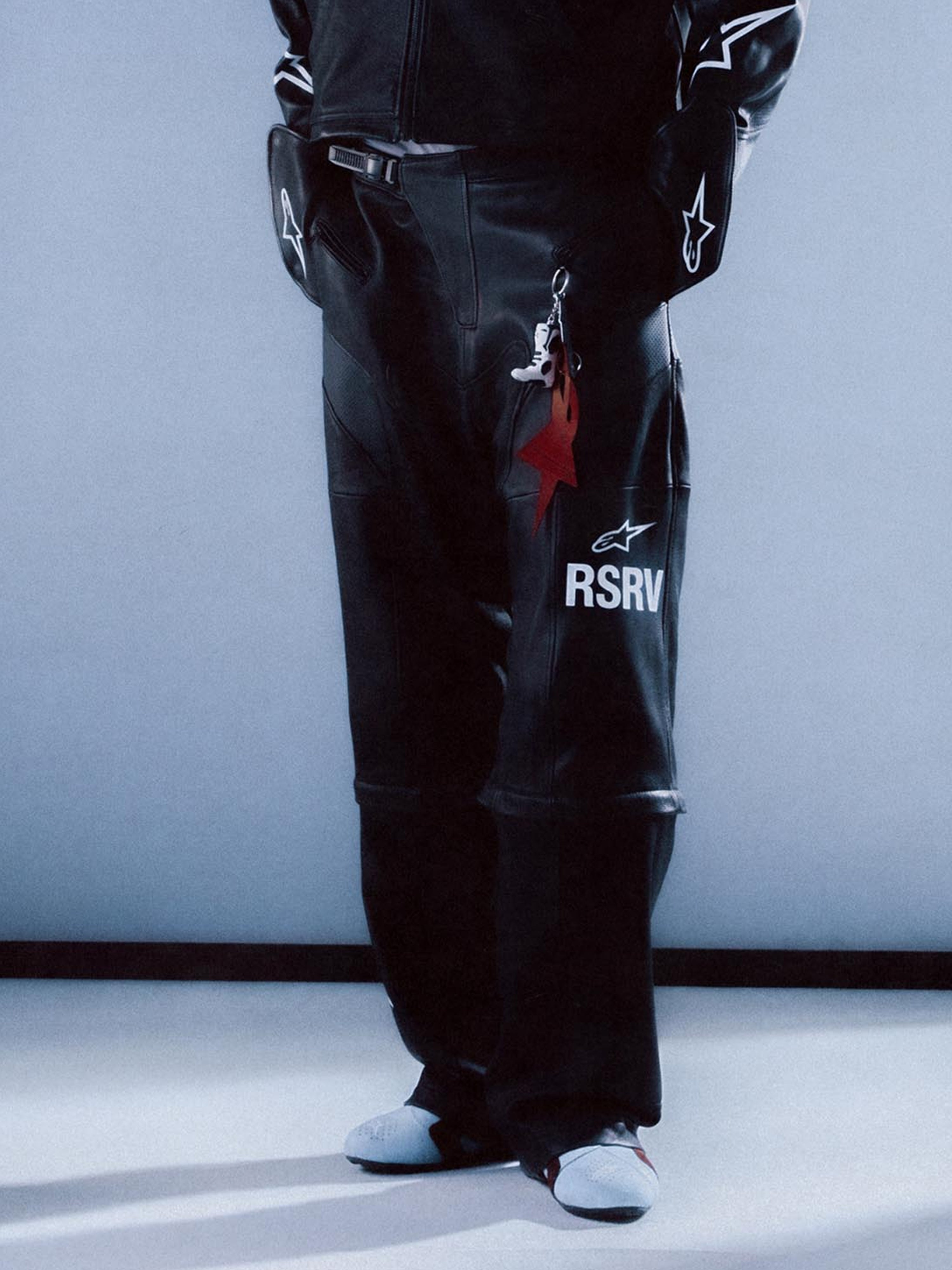 Alpinestars RSRV Neo Bat Leather Pants, pantaloni in pelle neri da moto streetwear, con logo RSRV bianco, dettagli stelle bianche, portachiavi stella rossa e stivale, outfit coordinato con giacca, scarpe bianche e nere