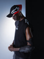 Alpinestars RSRV Tech Tank, canotta tecnica senza maniche nera con logo bianco sulla spalla, indossata con cappellino nero e rosso e gomitiera protettiva nera testurizzata