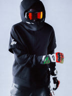 Alpinestars RSRV Vault Hoodie, felpa nera con cappuccio, stile streetwear motorsport, persona con casco e visiera arancione riflettente, guanti colorati rossi verdi e bianchi, design urbano premium