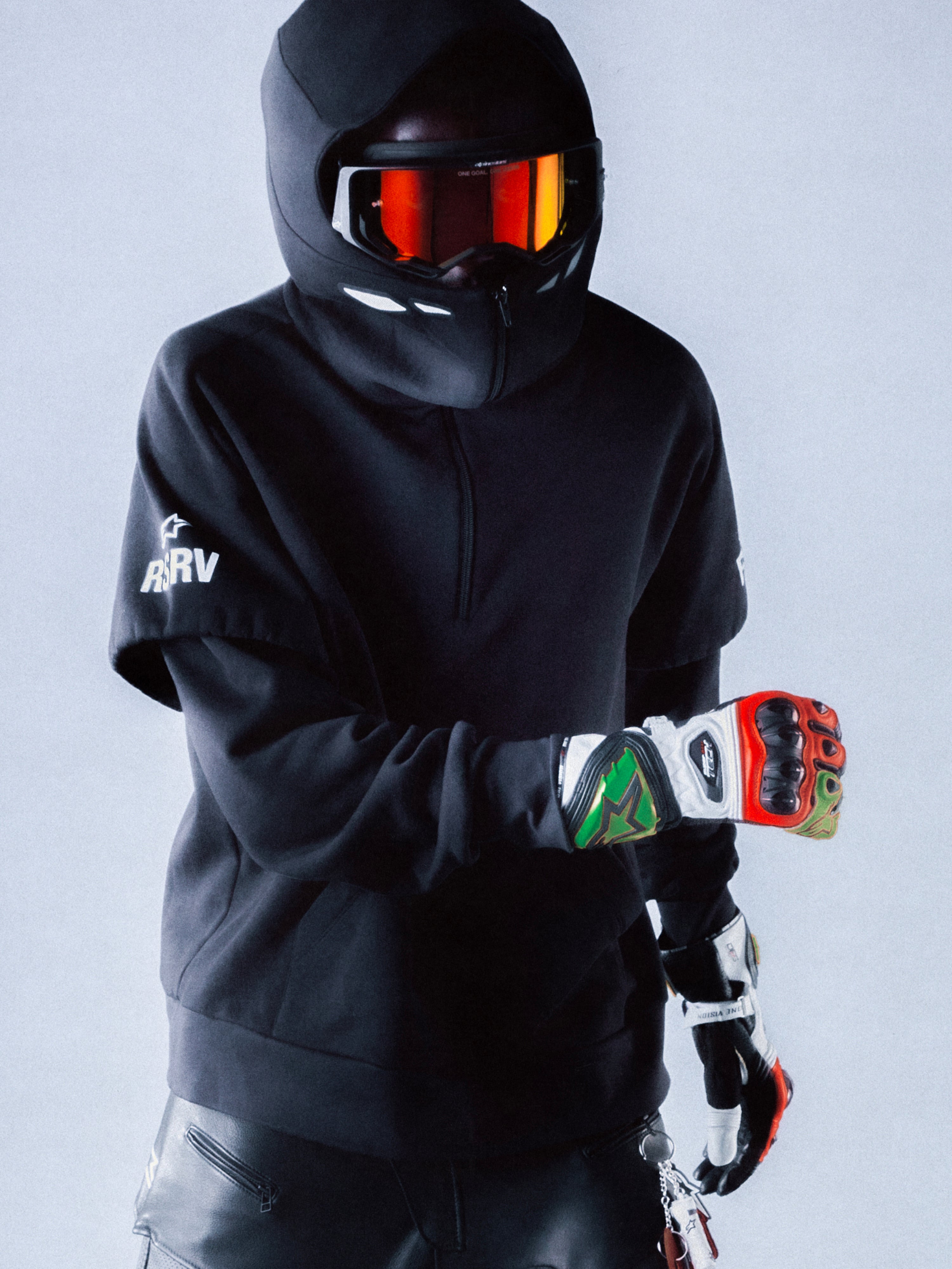 Alpinestars RSRV Vault Hoodie, felpa nera con cappuccio, stile streetwear motorsport, persona con casco e visiera arancione riflettente, guanti colorati rossi verdi e bianchi, design urbano premium
