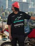 Alpinestars Aplomb Oversized CSF Tee, t-shirt a maniche corte, nero, con grande logo Astars sfumato verde acqua e bianco e grafica con bandiera a scacchi, indossata con un berretto corallo, in piedi accanto a una motocicletta rossa, streetwear casual lifestyle